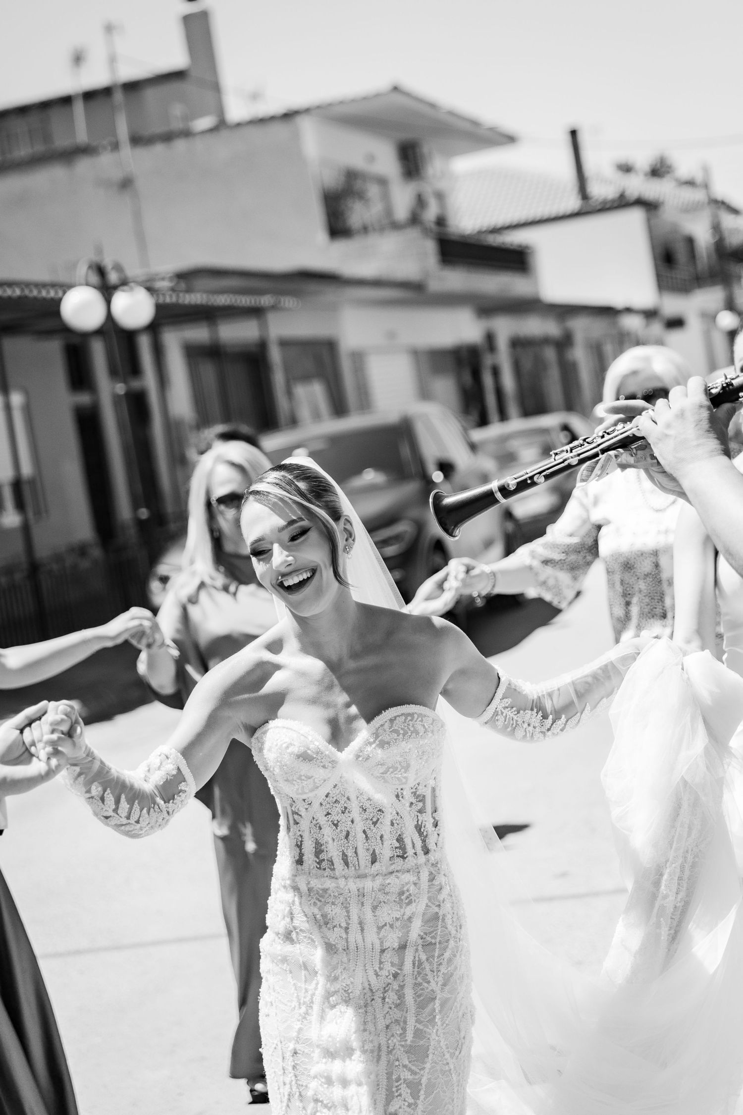 Black and white photo of a smiling bride in lace gown dancing outdoors with guests and a clarinet player at her wedding.