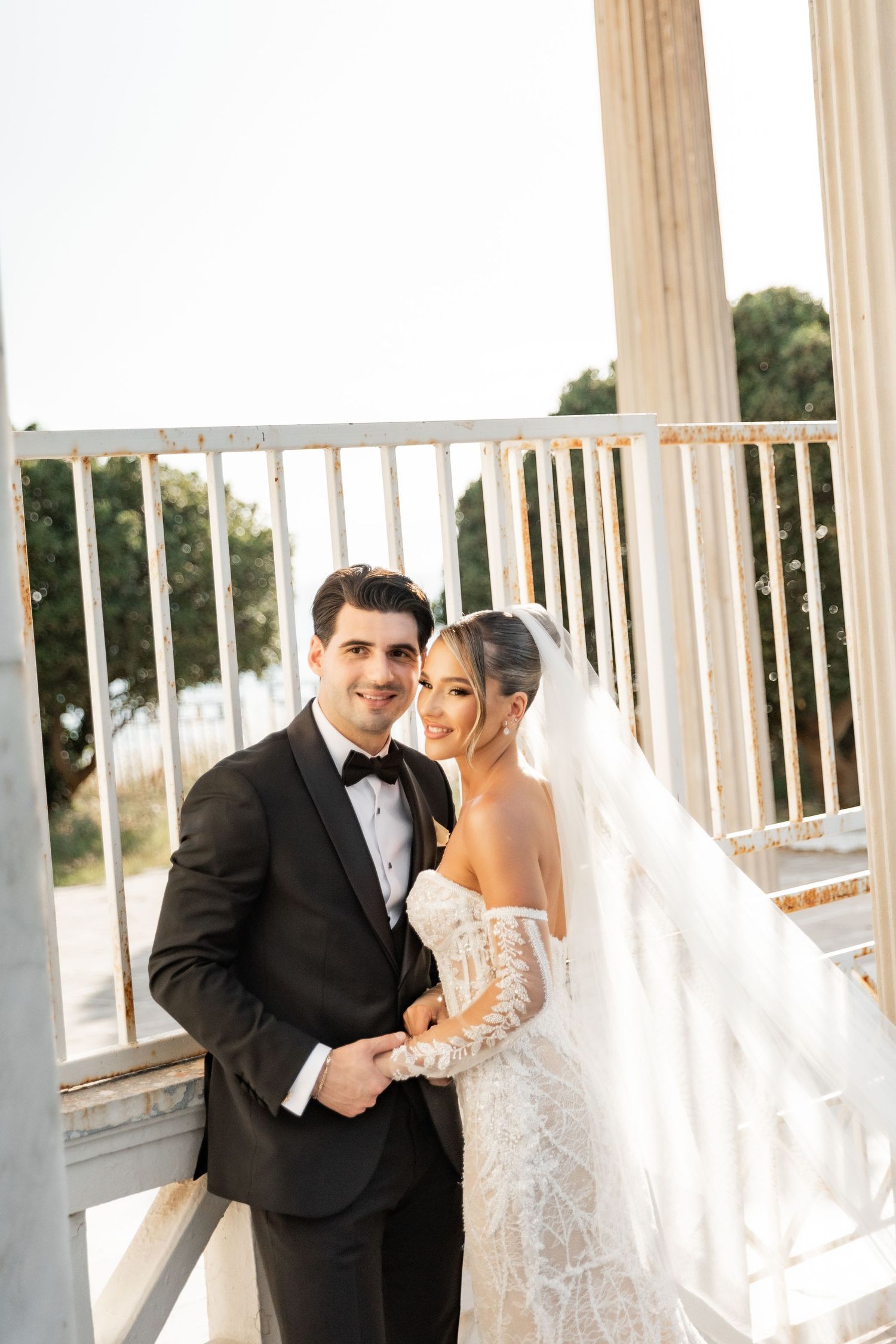 A bride in a lace gown and veil poses with a groom in a black tuxedo near elegant white columns outdoors.