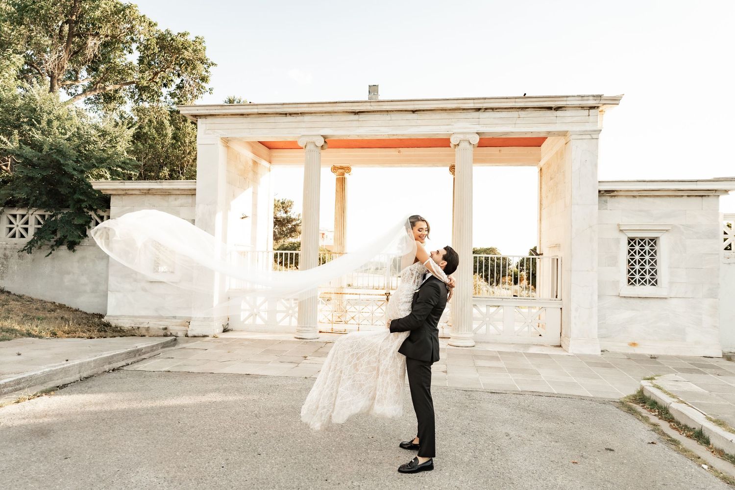 Groom lifting bride in wedding attire before a white columned villa at sunset.