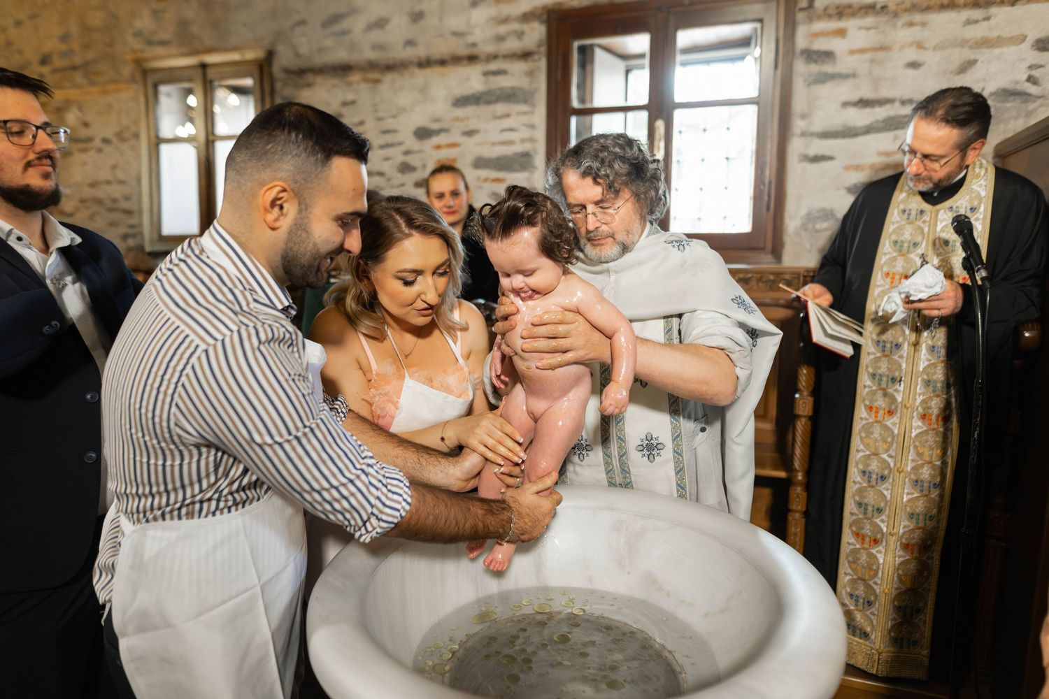 A baptism ceremony taking place at a church with a priest and family gathered around a baptismal font.