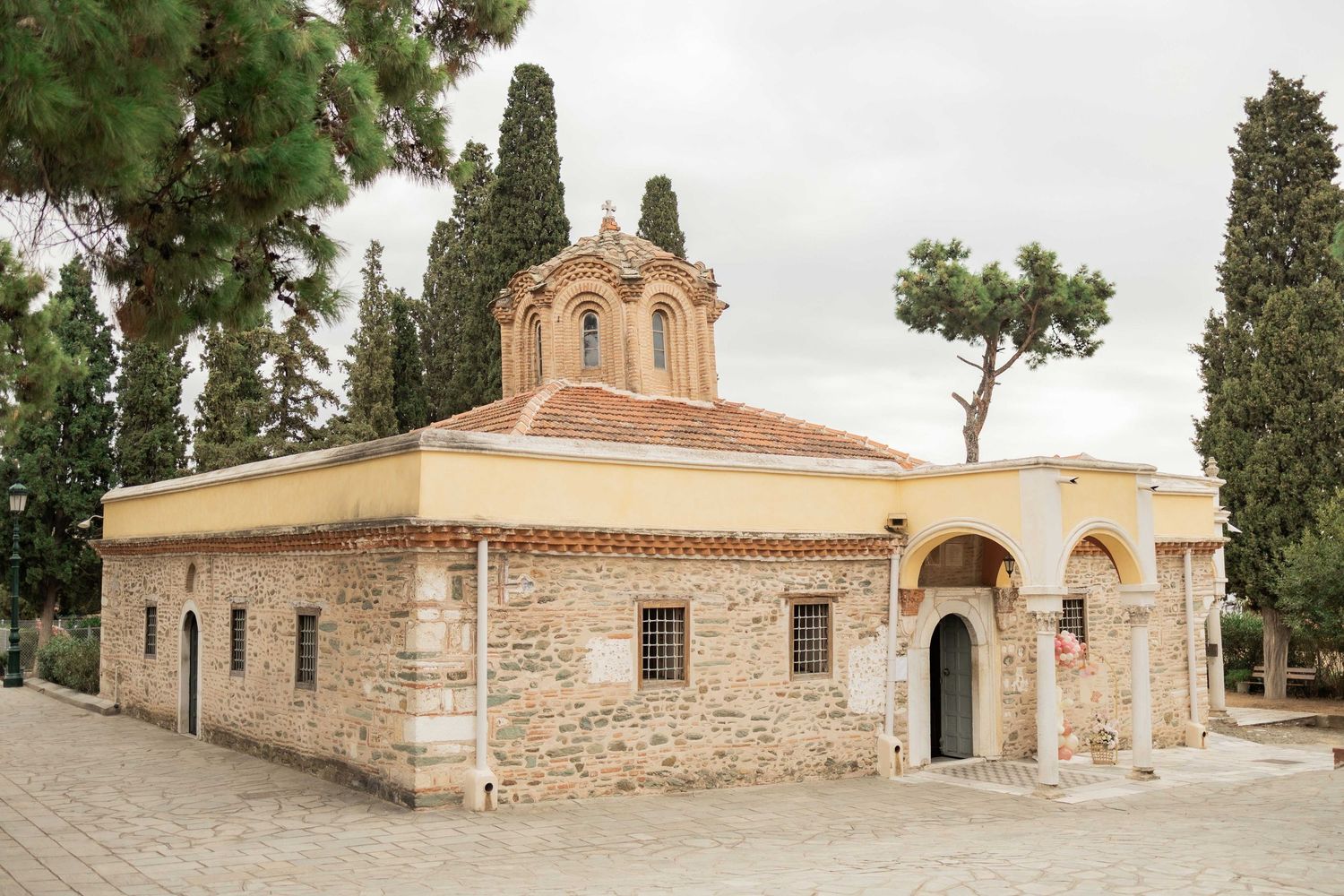 A panoramic view of a traditional Greek Orthodox monastery with stone buildings and cypress trees against a cloudy sky.