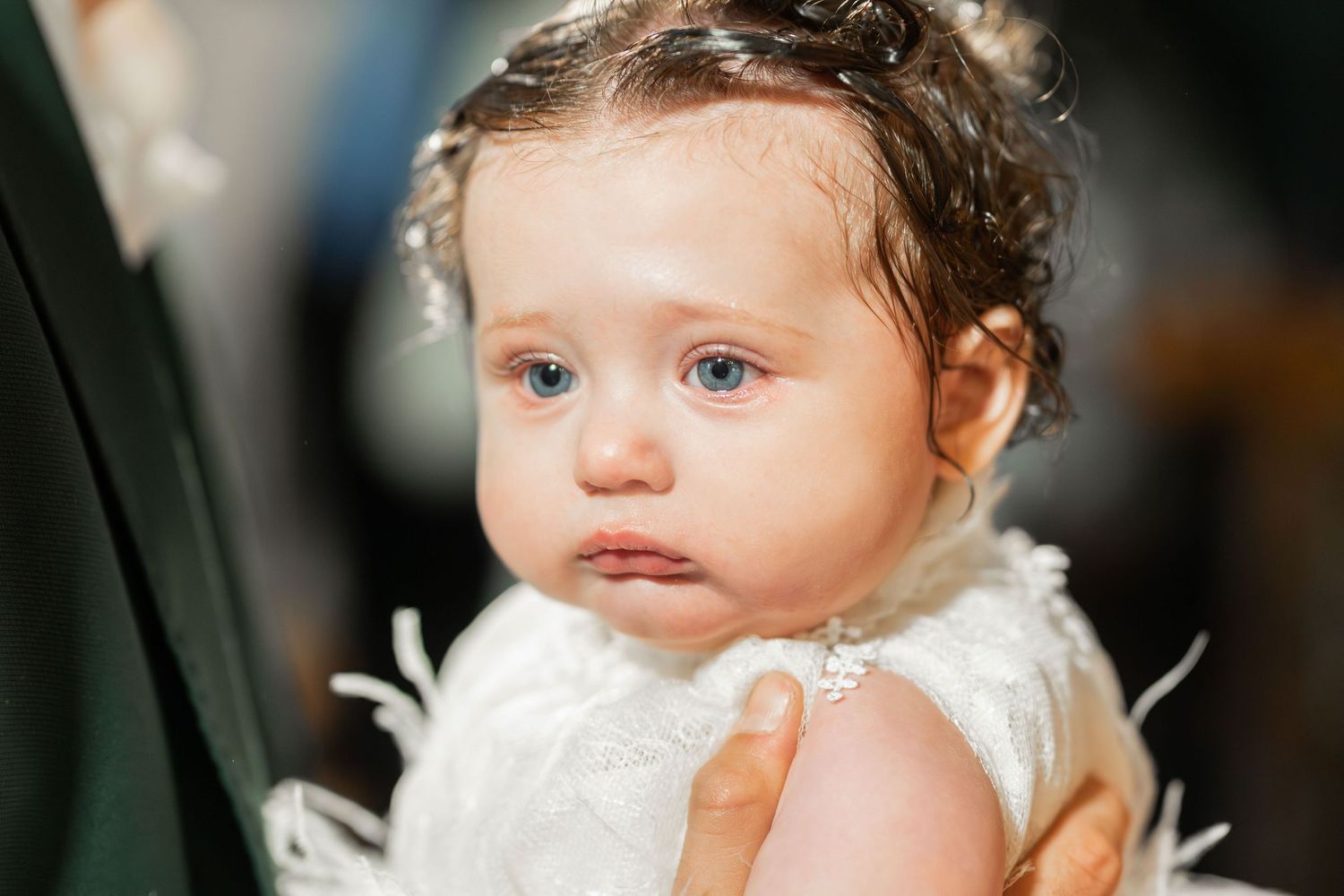 A small child wearing a white lace dress with delicate details looks off to the side with rosy cheeks.