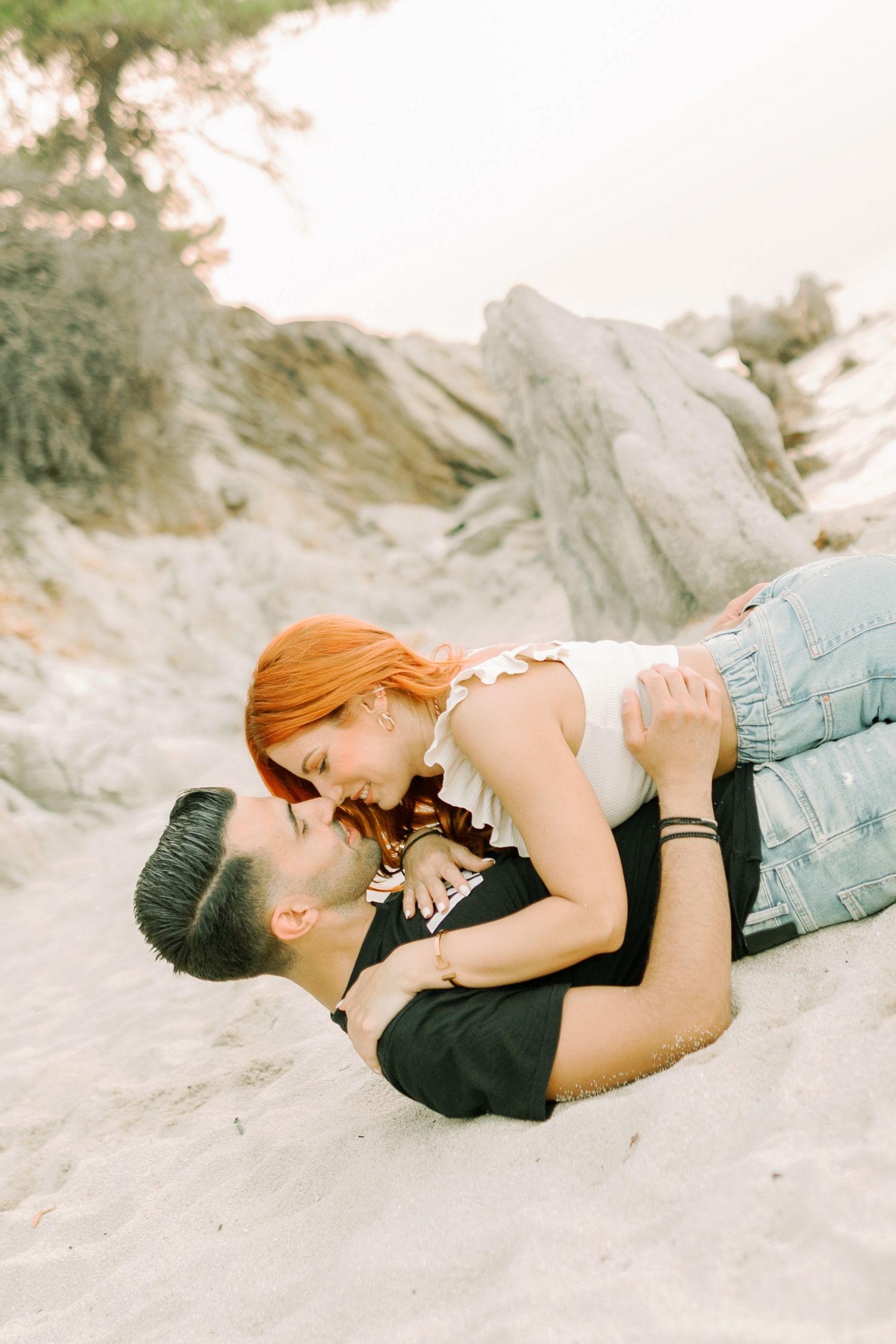 A romantic couple lying together on a sandy beach with rocks and greenery in the background.