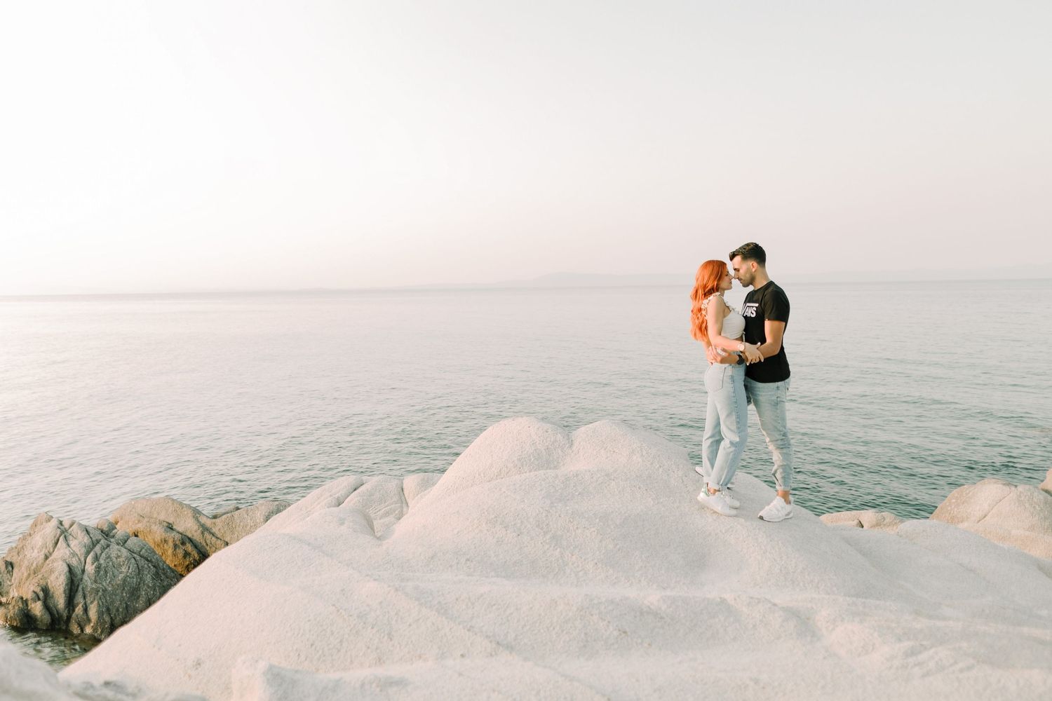 A couple embraces while standing on snow-covered rocks overlooking a misty ocean at sunset.