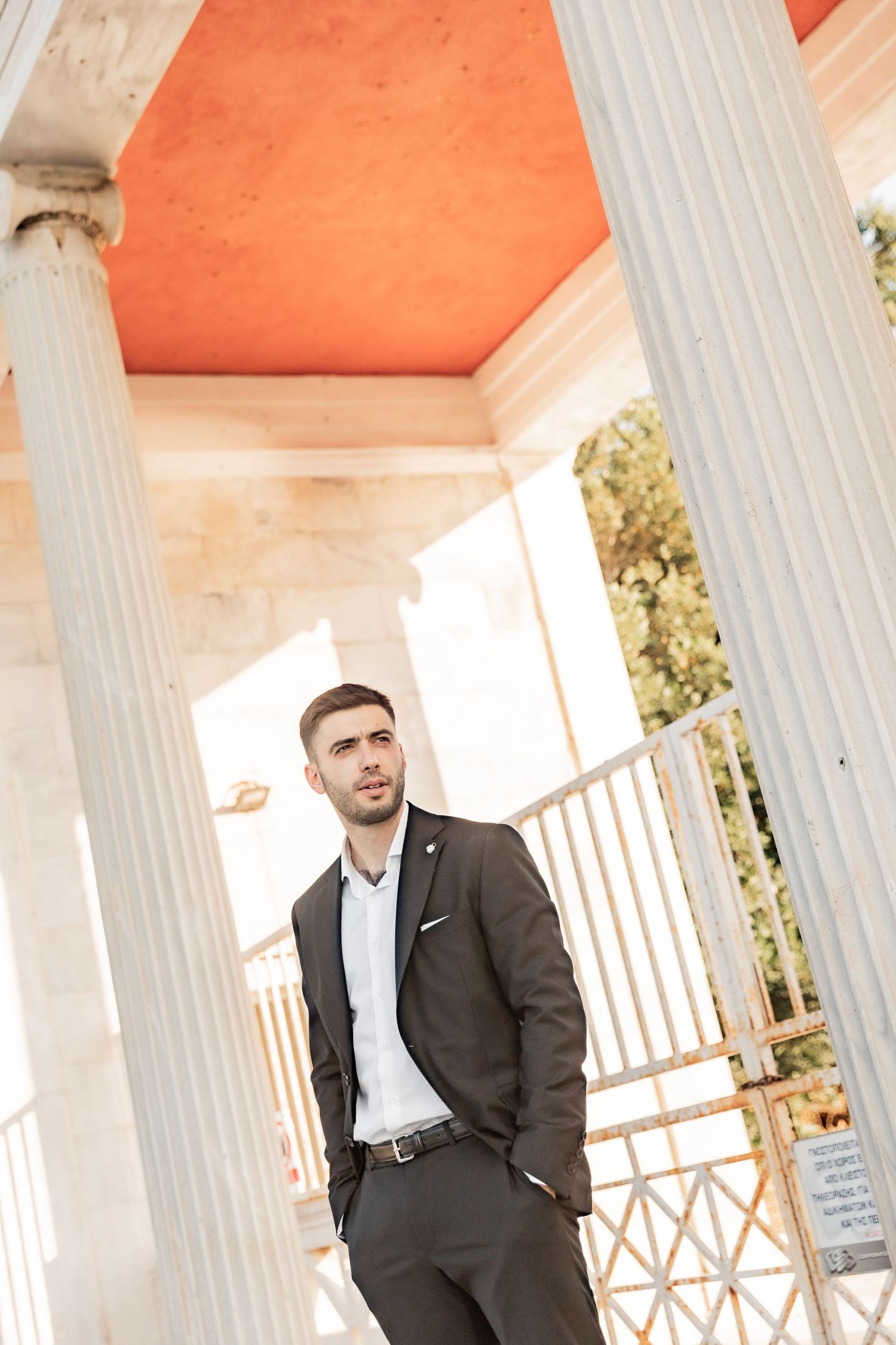 A well-dressed man in a dark suit stands confidently between white classical columns with an orange ceiling.