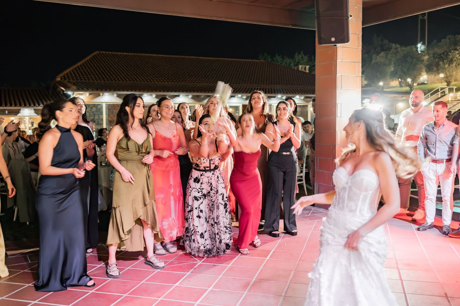 Bride tossing bouquet to excited wedding guests in colorful dresses at an outdoor evening reception.