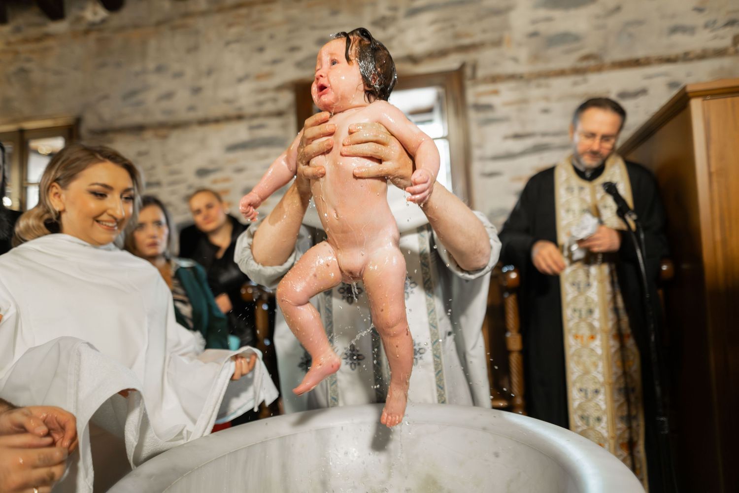 A newborn baby is being lifted up during a special ceremony or celebration in a rustic stone-walled room.