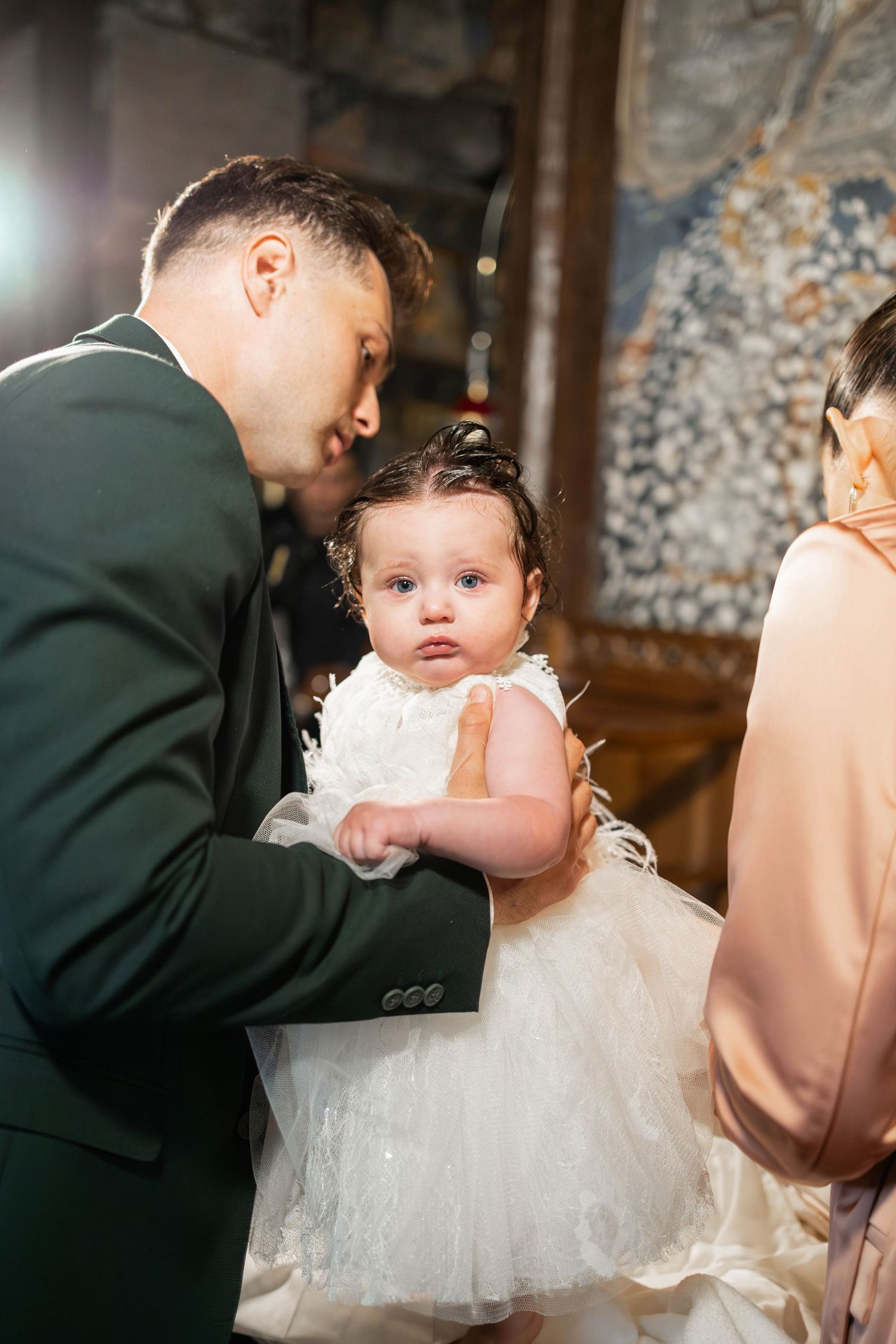 A baby in a white tulle dress is being held during what appears to be a wedding ceremony.