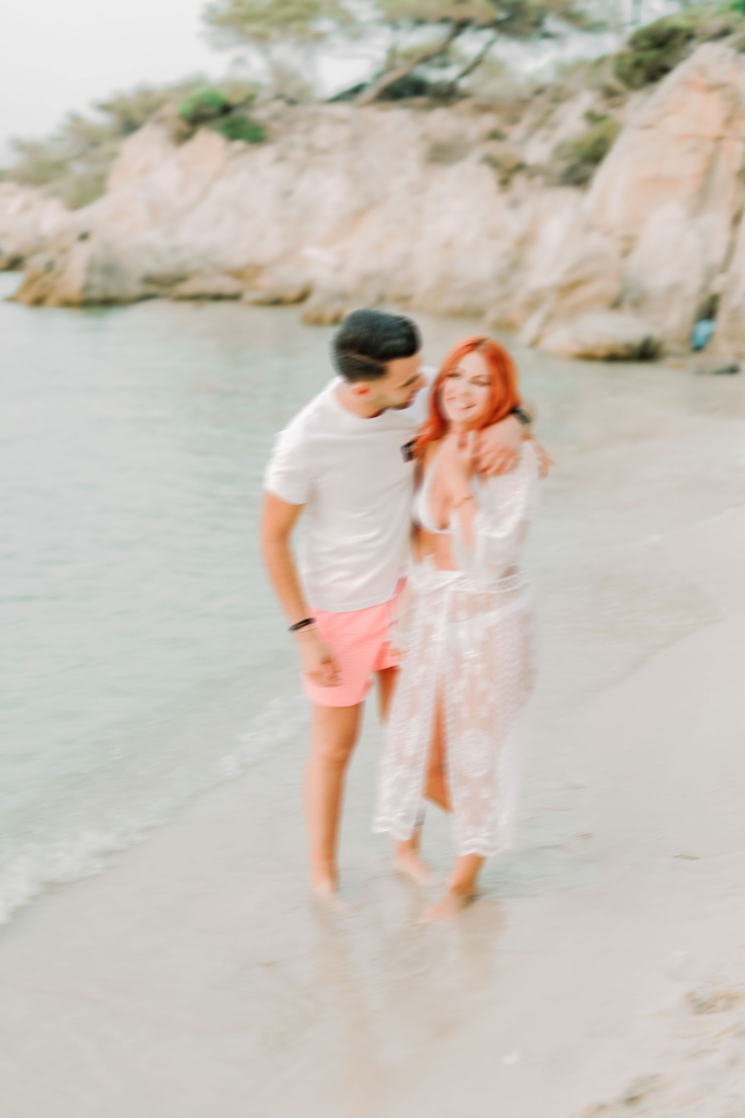 A couple walks along a scenic beach shoreline with rocky cliffs in the background.