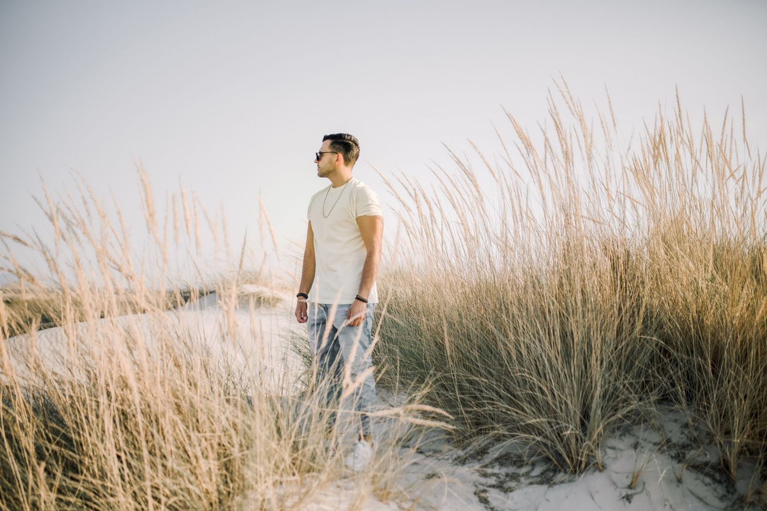 Person in white clothing walks through tall golden beach grass during sunset along the coastline.