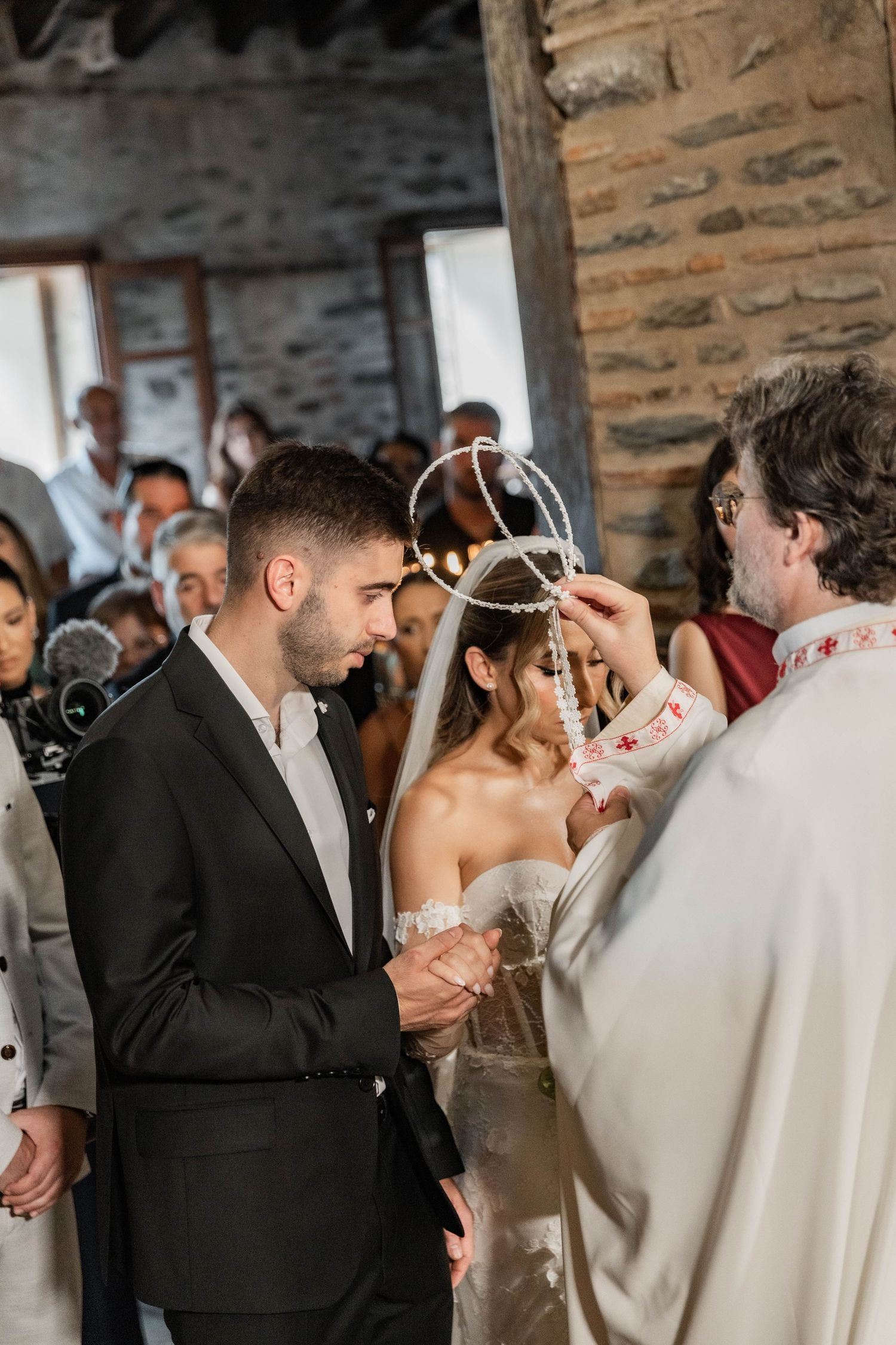 Orthodox wedding ceremony with priest placing crowns on bride and groom in rustic stone church.