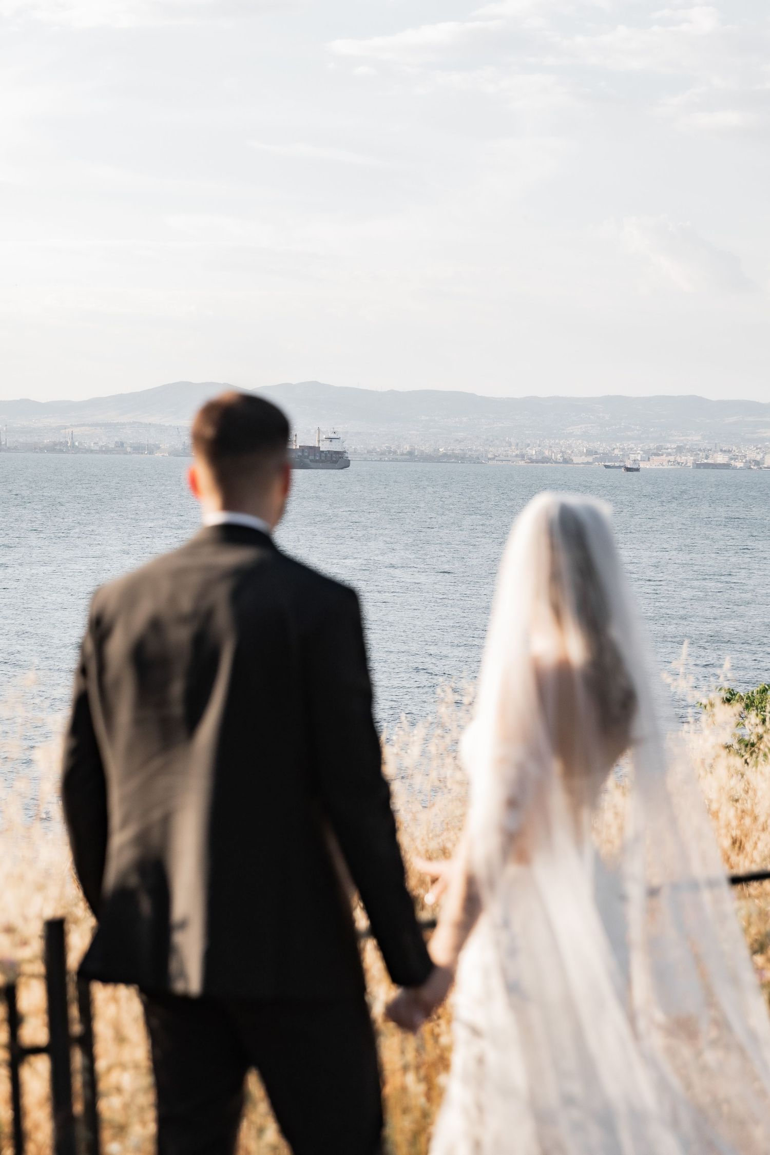 Bride and groom holding hands overlooking a scenic sea view with mountains and a ship in the background.