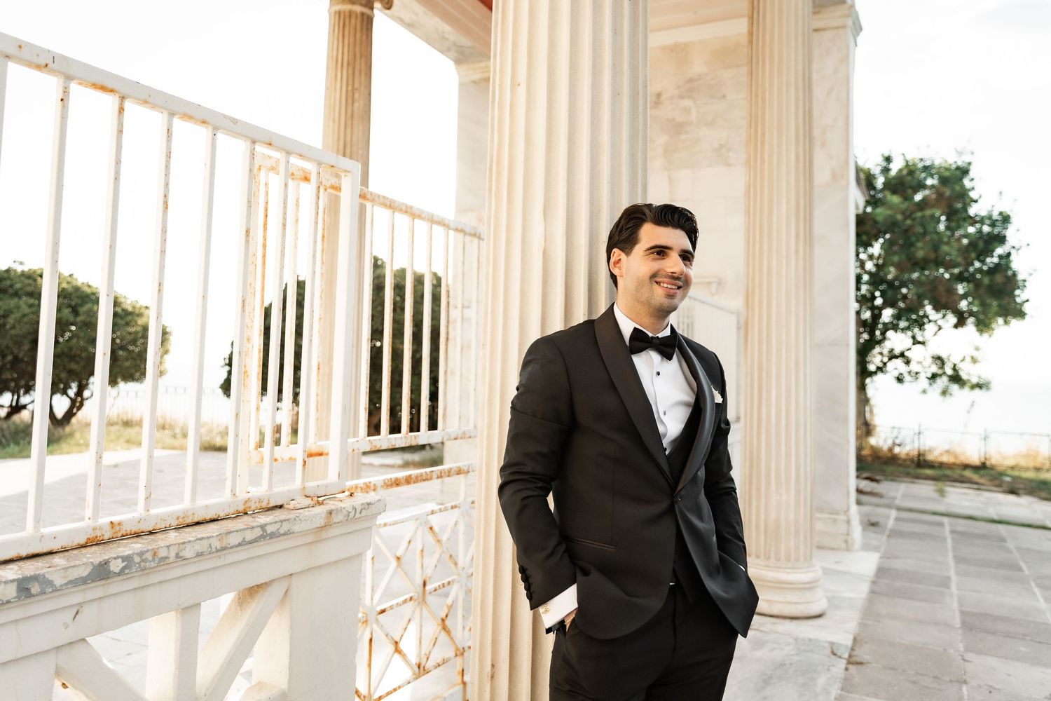 Groom in black tuxedo with bow tie stands confidently between white marble columns at elegant outdoor wedding venue.