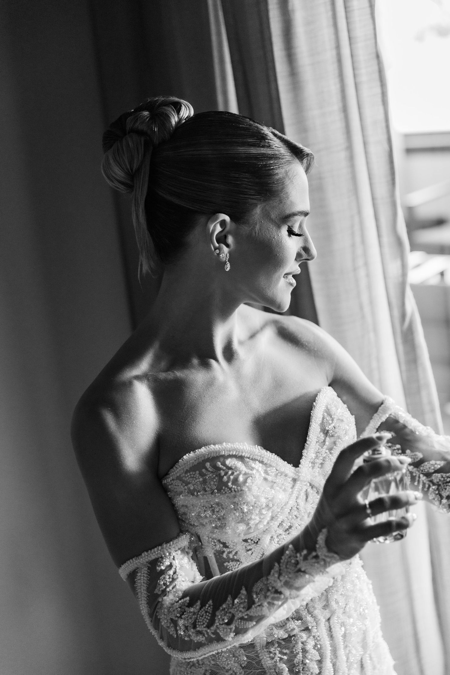 Black and white photo of a bride in a lace strapless gown, smiling near a window while adjusting her dress.