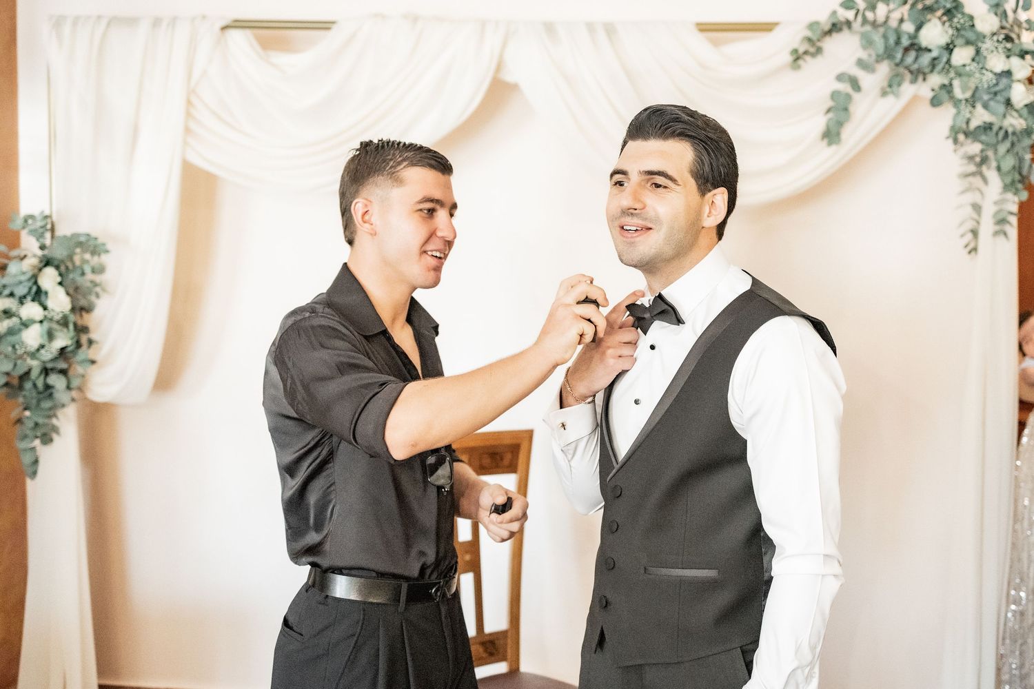 A groomsman adjusts the bow tie of a groom in a vest at a wedding venue decorated with white drapes.