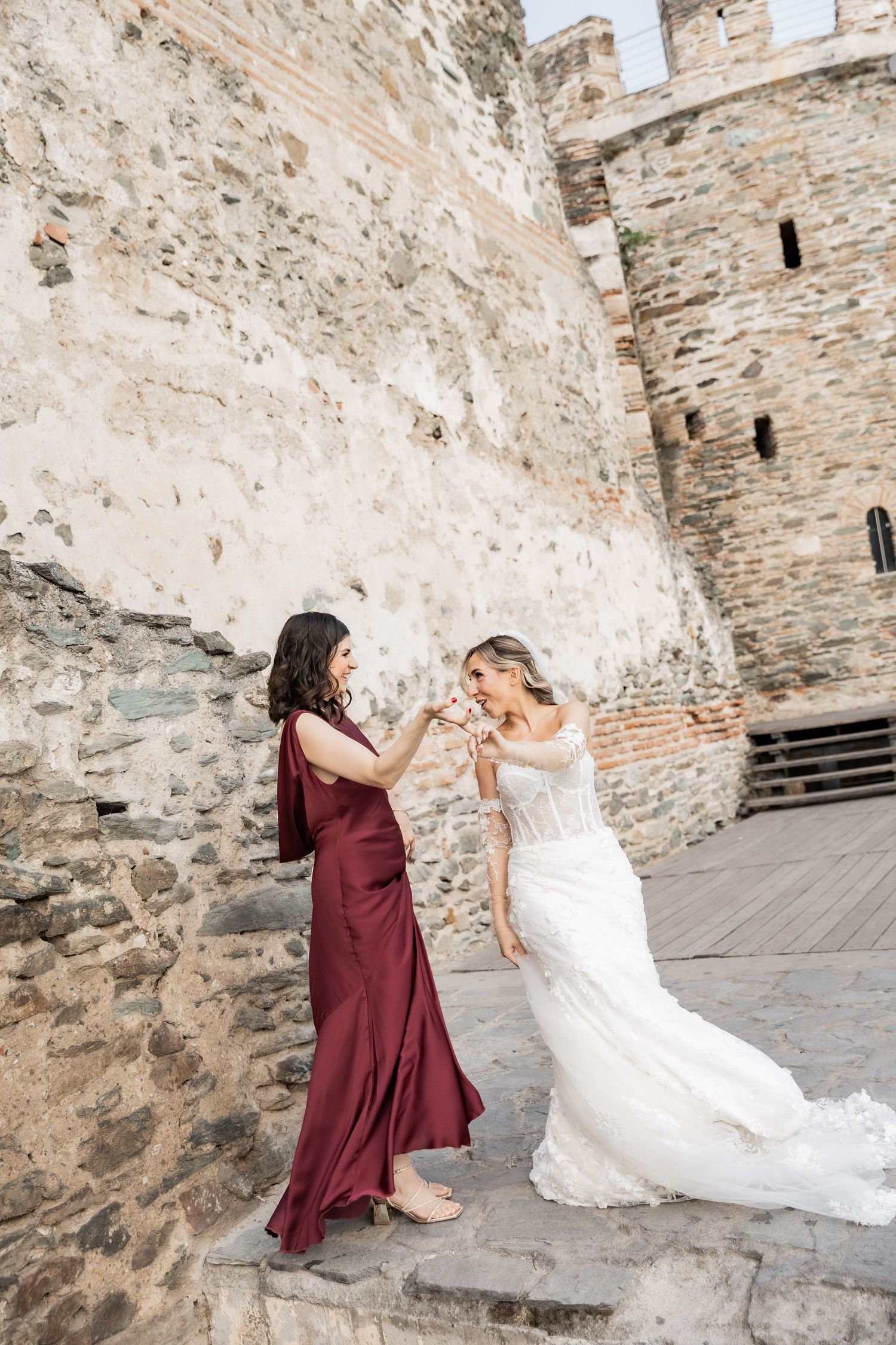 Bride in white gown and bridesmaid in burgundy dress toasting champagne on stone steps of historic castle.