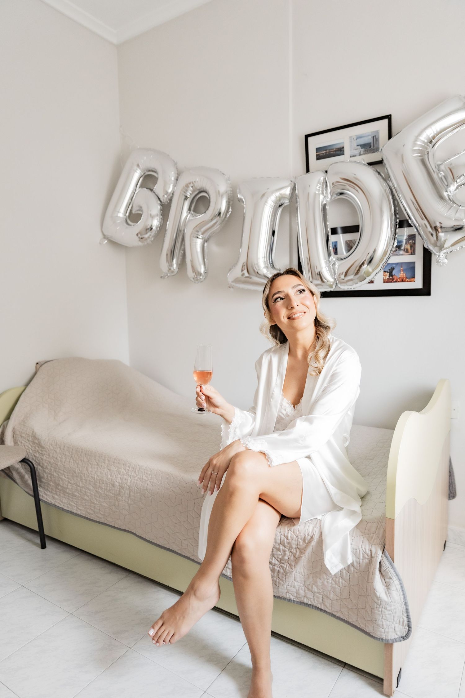A smiling bride in a white robe sits on a bed holding a champagne glass, with silver BRIDE balloons on the wall.