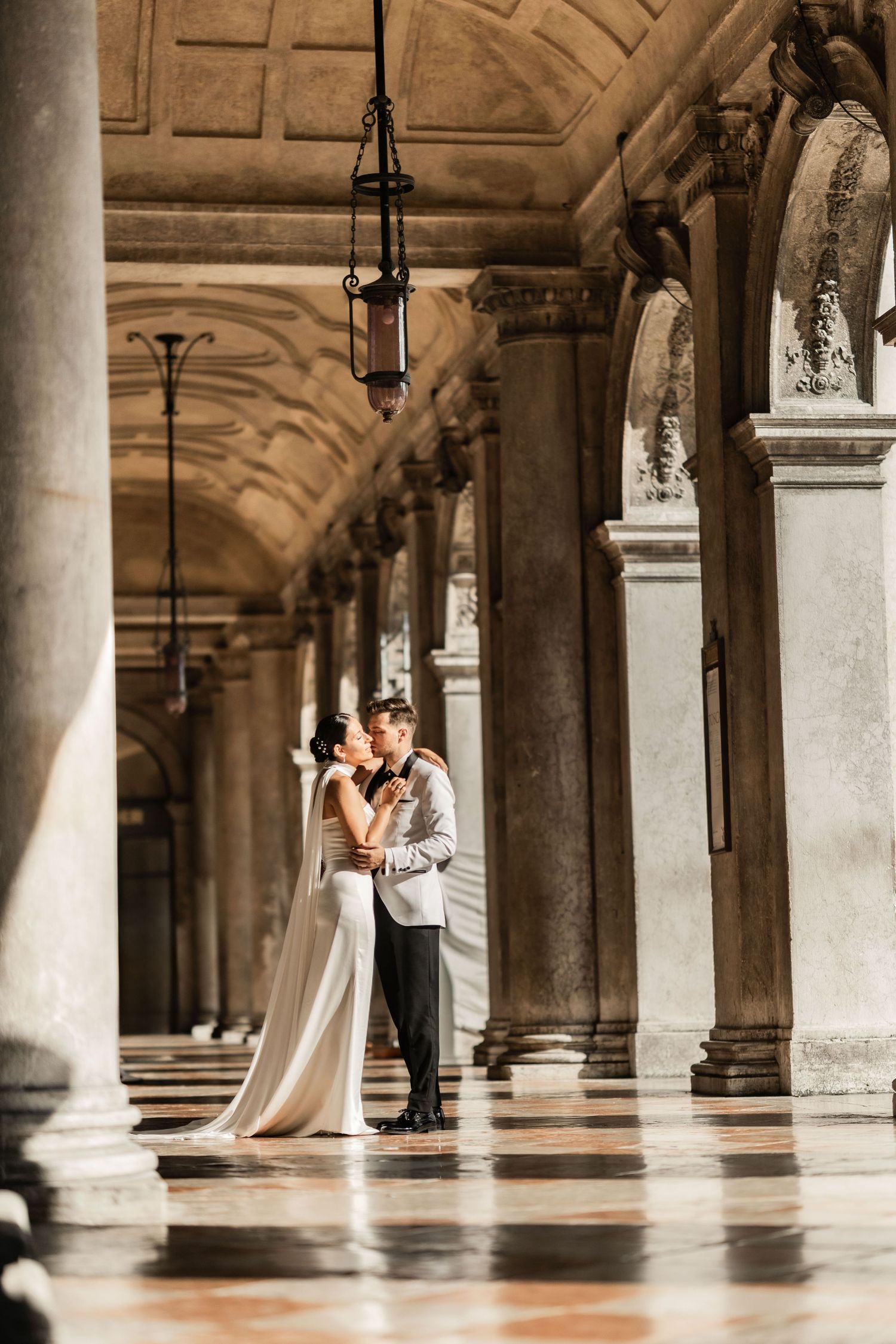 A romantic moment captured in a historic Venetian colonnade with ornate stone columns and vaulted ceilings.