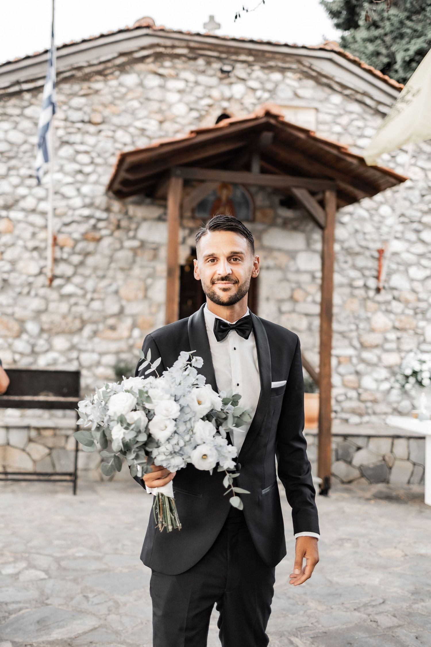 A person in formal attire holding a white floral bouquet against a rustic stone wall backdrop.