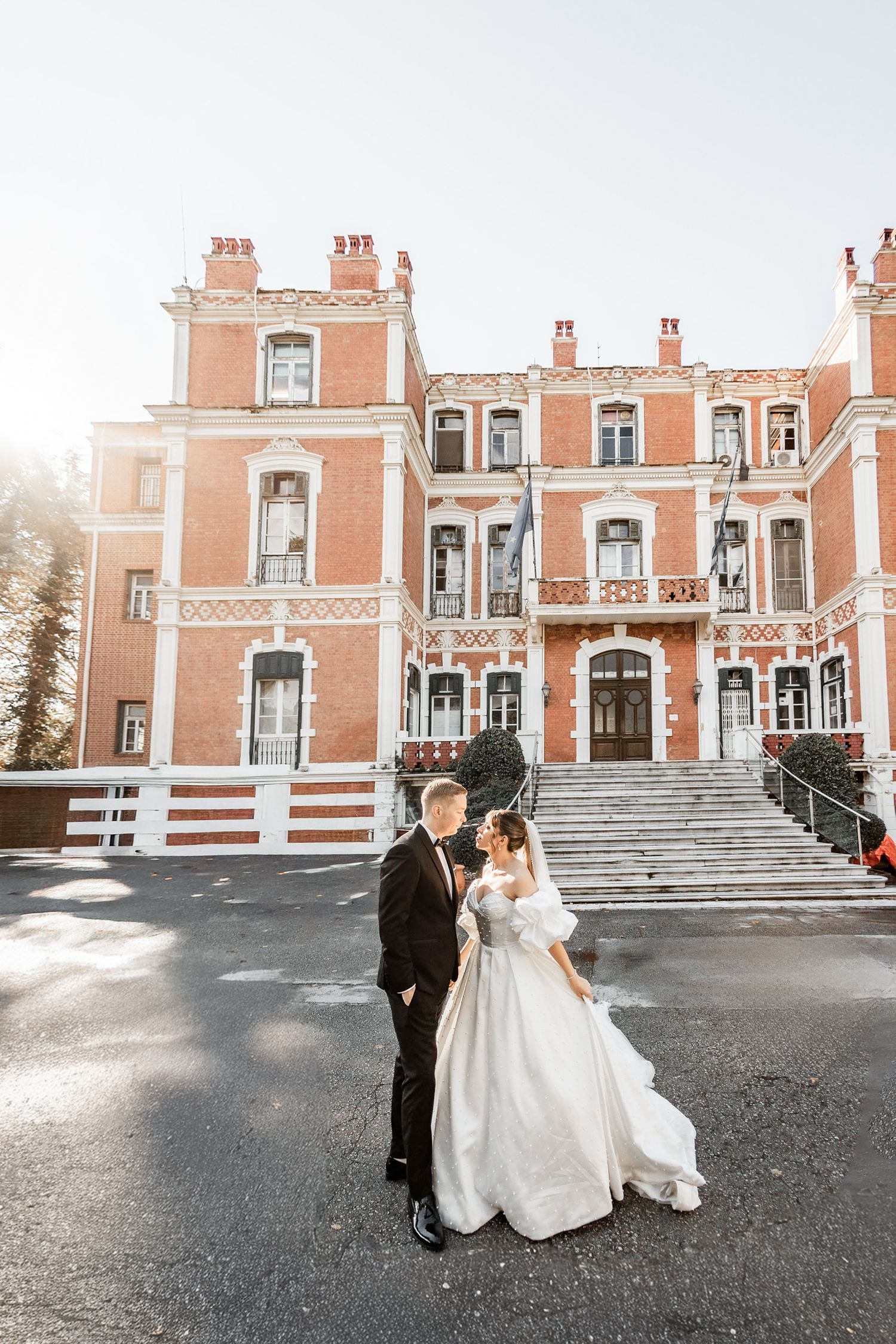 Elegant newlyweds pose in front of a grand coral-colored mansion with ornate architecture.