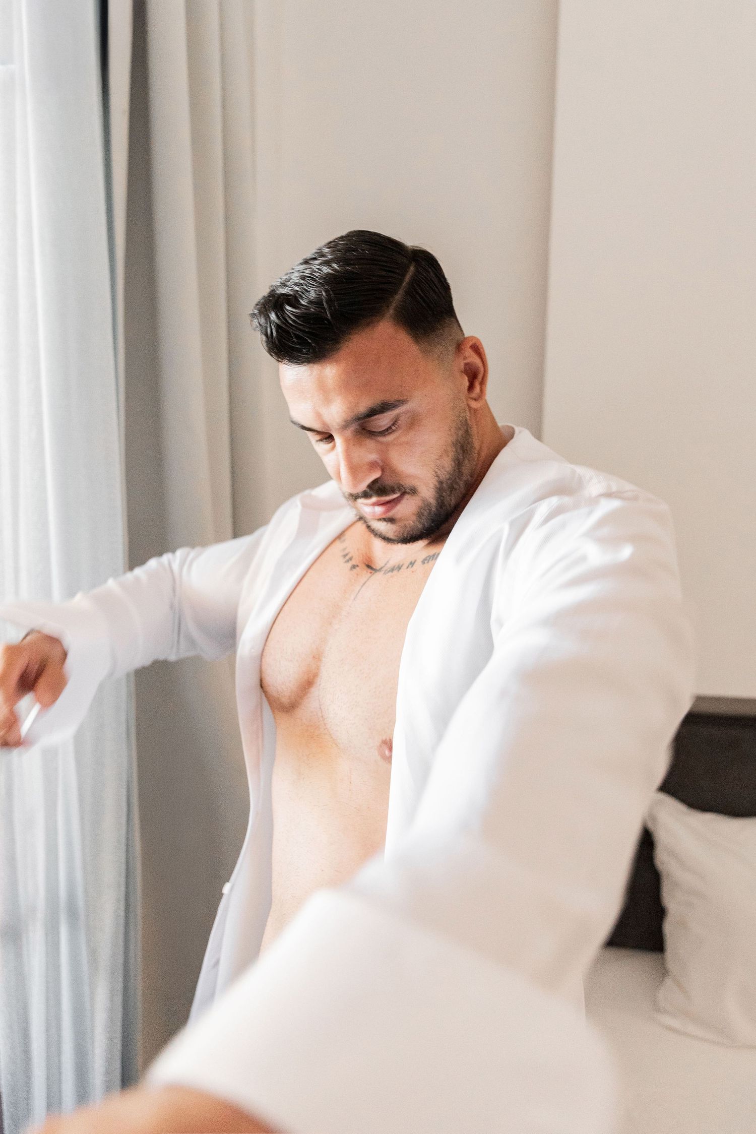 Groom getting dressed in a white button-up shirt against white curtains.