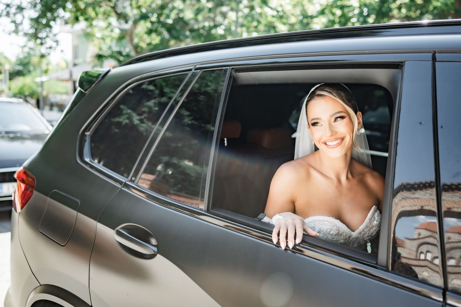 Smiling bride in wedding dress and veil leaning out of a dark luxury car window on her wedding day.