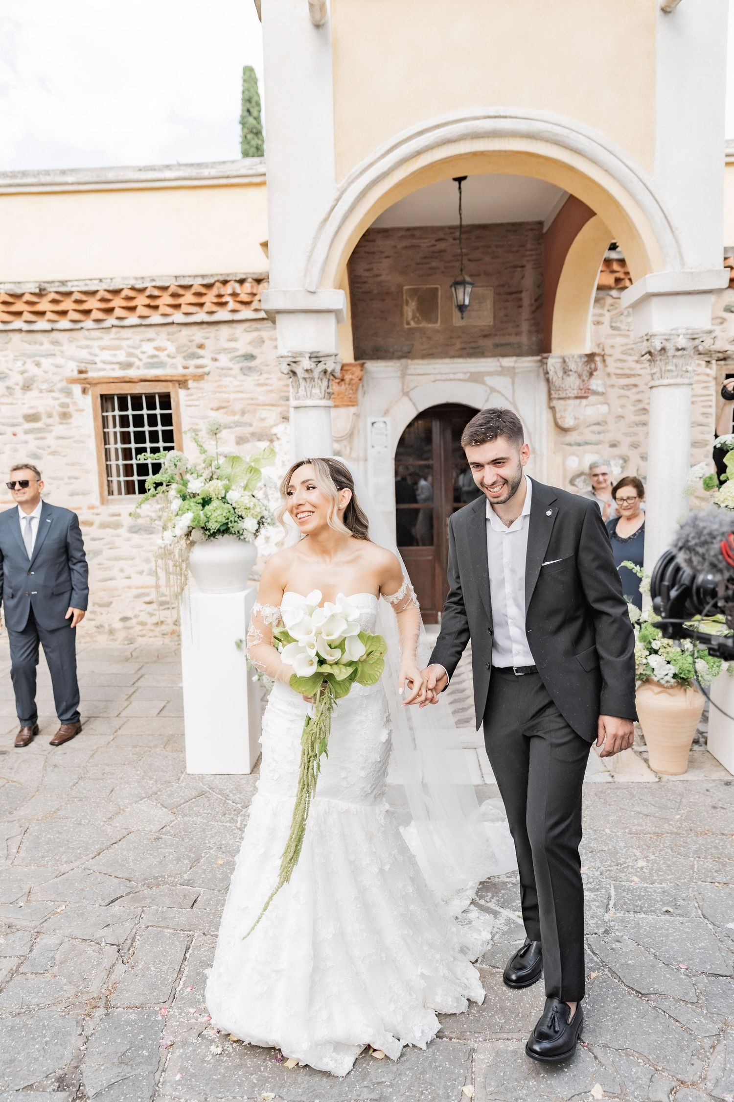 Bride and groom holding hands, smiling as they exit a rustic stone church, bride in lace mermaid gown holding white bouquet.