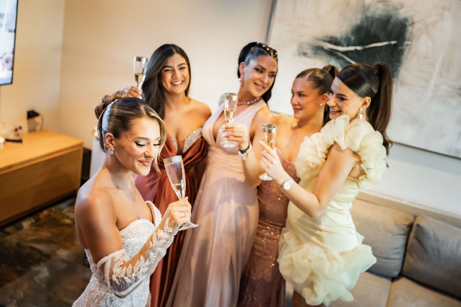 Bride and bridesmaids in elegant gowns toasting with champagne glasses during wedding preparations.