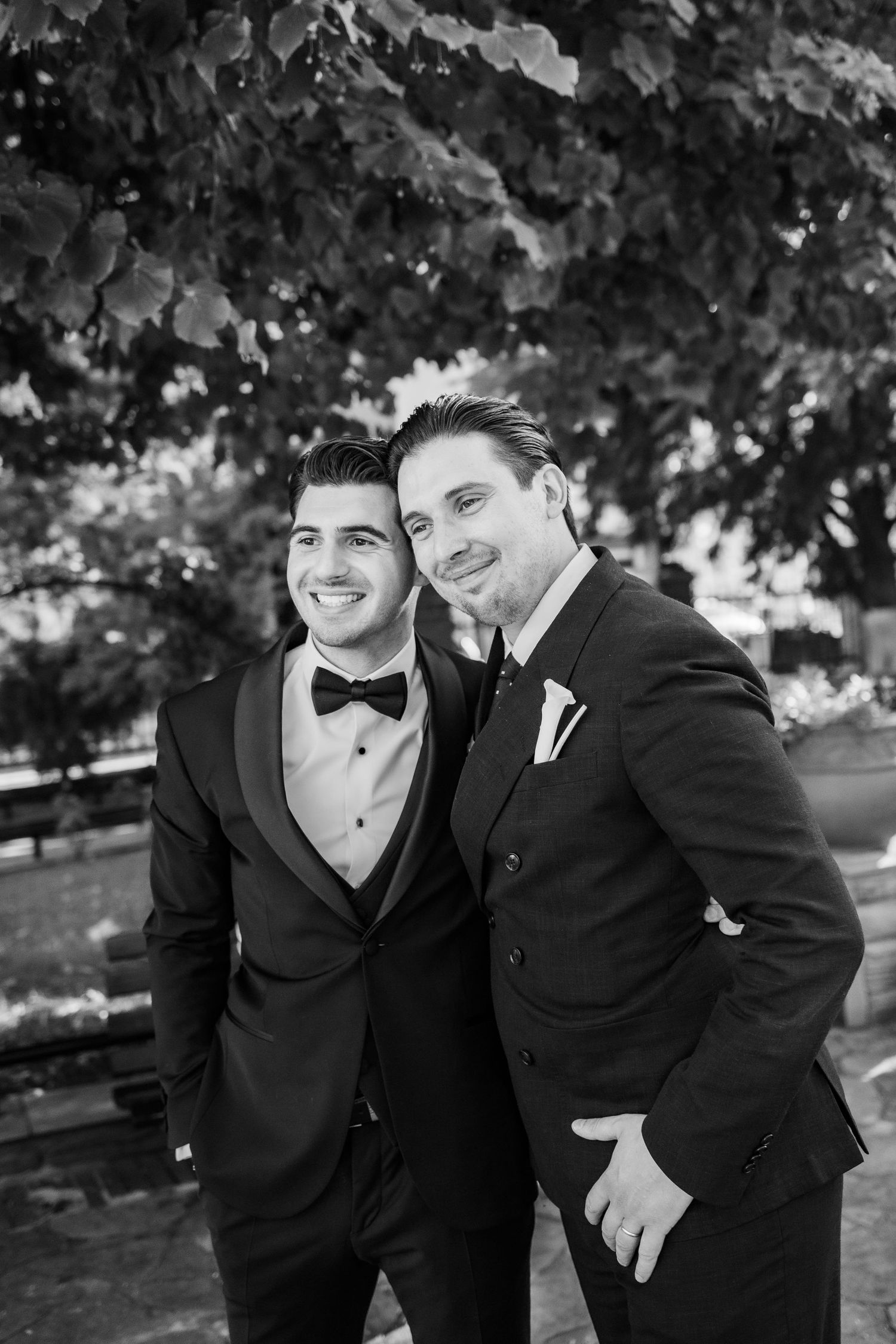 Two grooms in black tuxedos smiling together outdoors on their wedding day, black and white photo.
