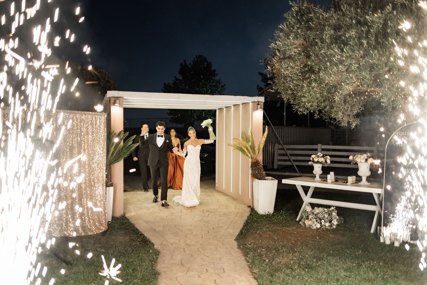 Wedding couple exits through sparkler tunnel at night with guests celebrating in decorated outdoor venue.