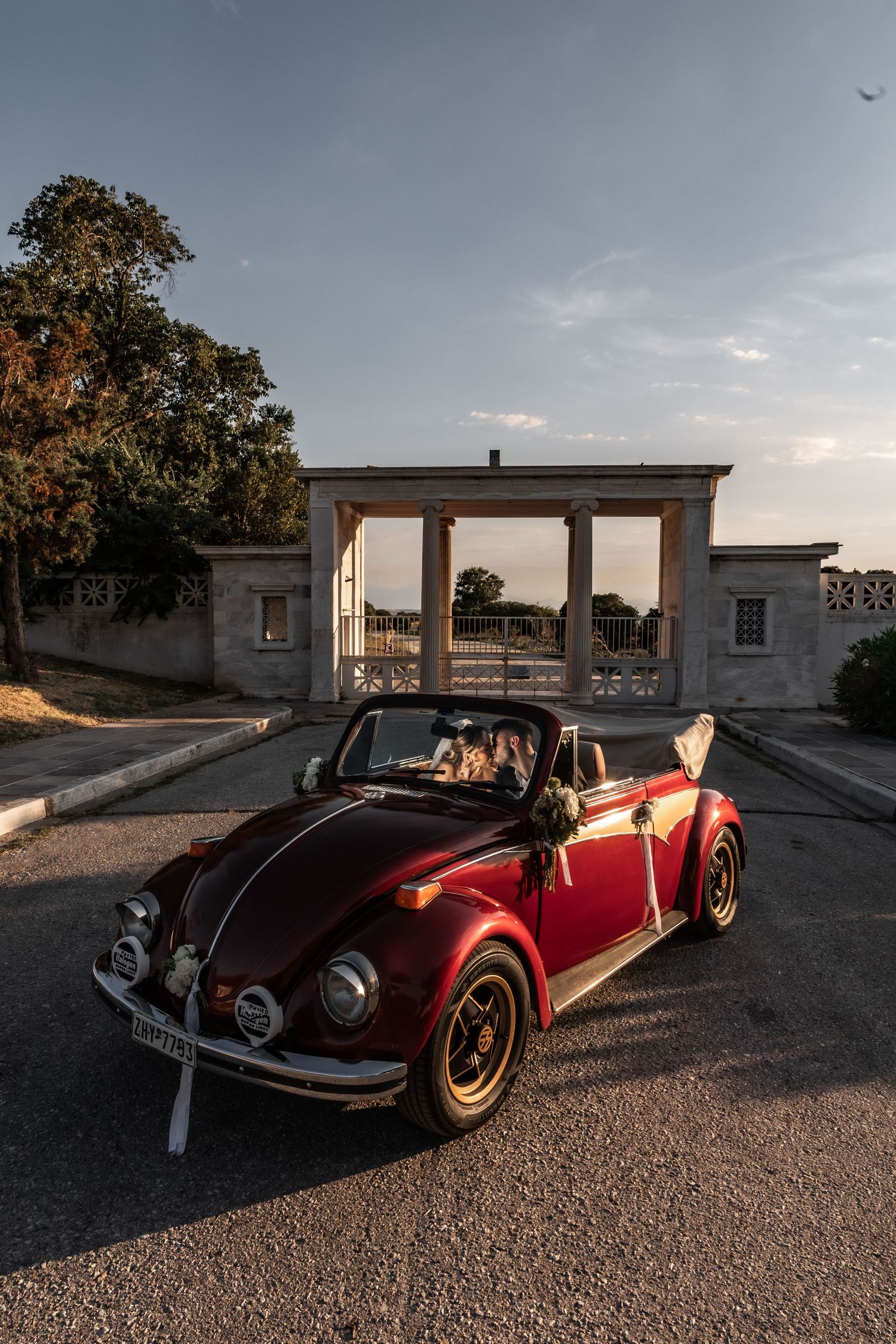 Red vintage VW Beetle convertible with wedding decorations parked at elegant venue gates during golden hour sunset.