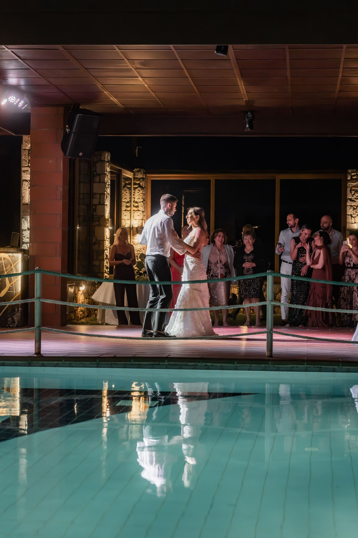 Bride and groom share their first dance beside an indoor pool while wedding guests watch in the background.
