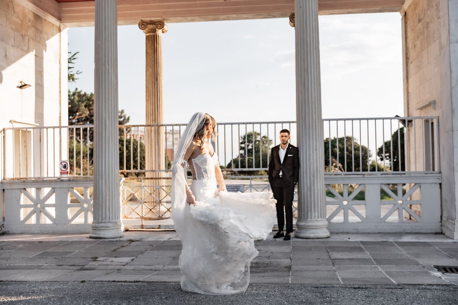 Bride in flowing white gown with veil stands before elegant columns as groom in black tuxedo looks on at outdoor wedding venue.