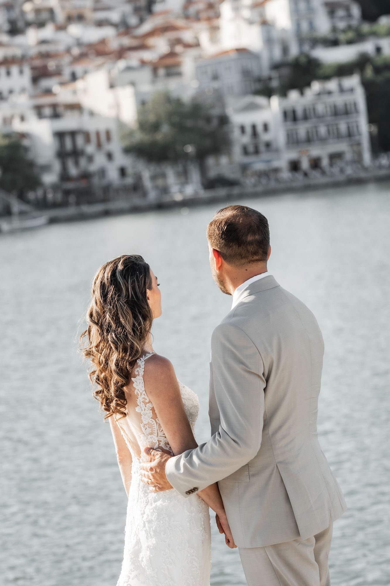 Wedding couple walks along waterfront with Mediterranean village backdrop.