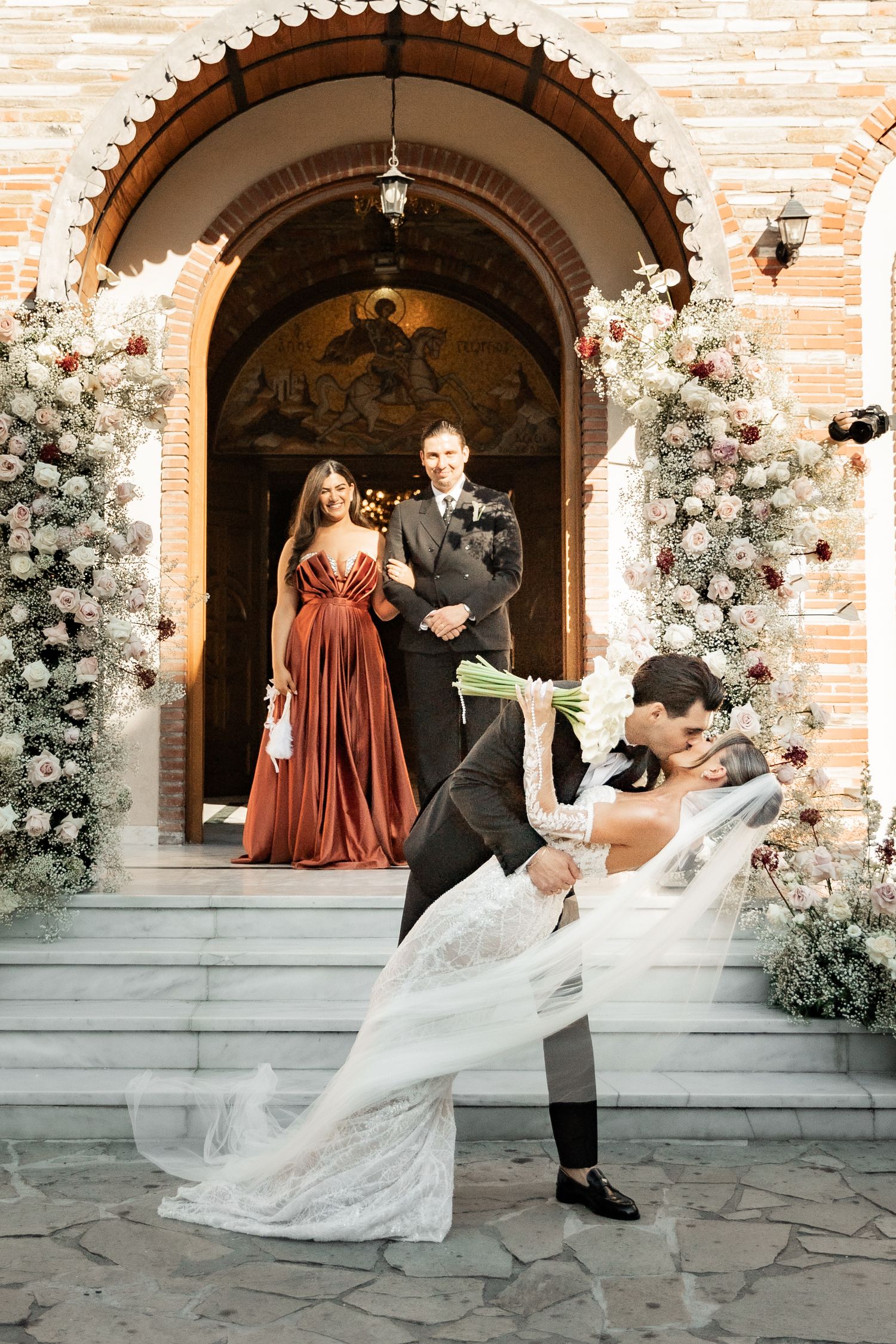 Groom dips bride in elegant kiss outside floral-adorned church entrance, with bridesmaid and groomsman watching behind.
