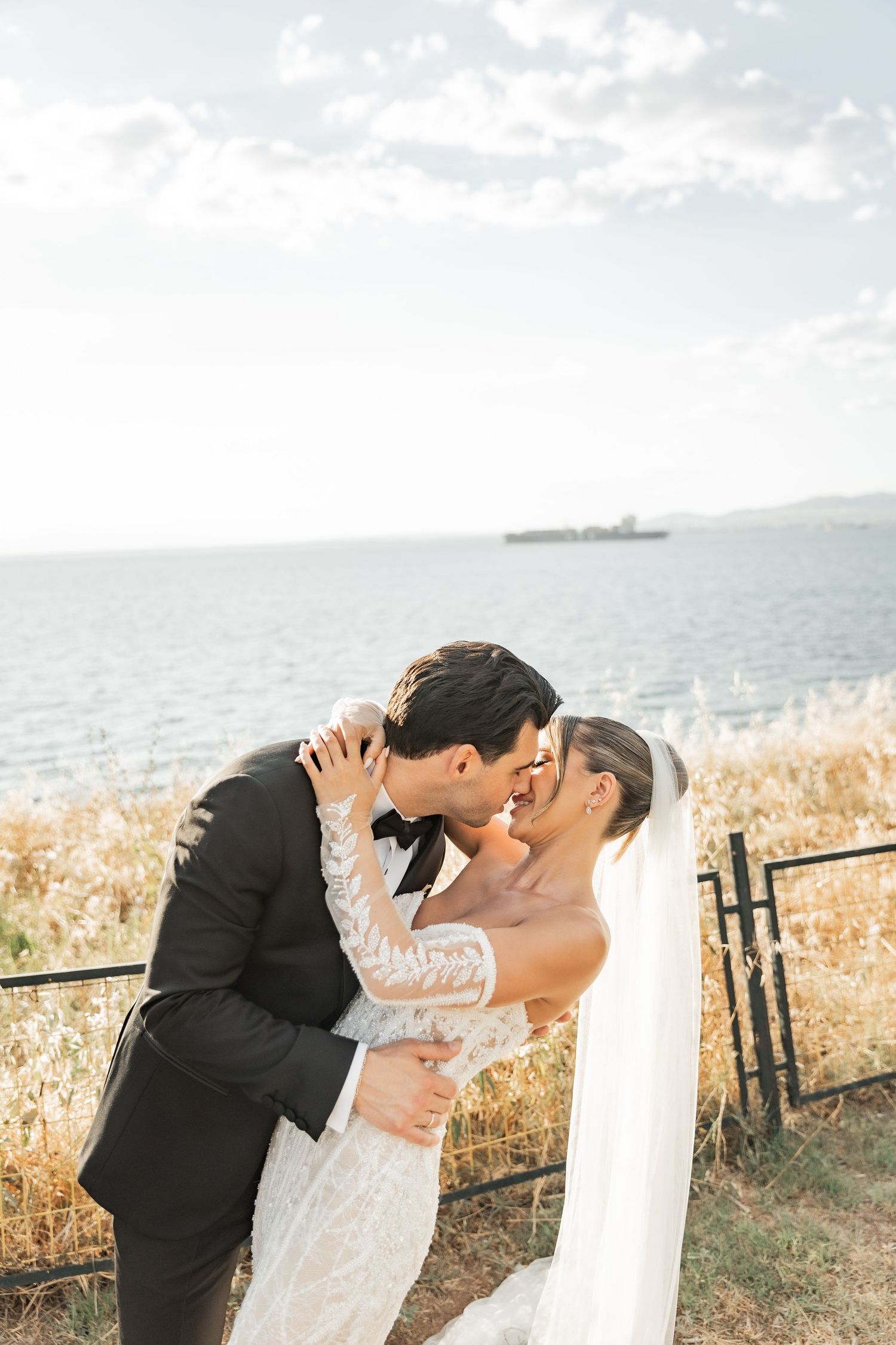 Bride and groom share a romantic kiss by the seaside, dressed in elegant wedding attire with a veil flowing in the breeze.