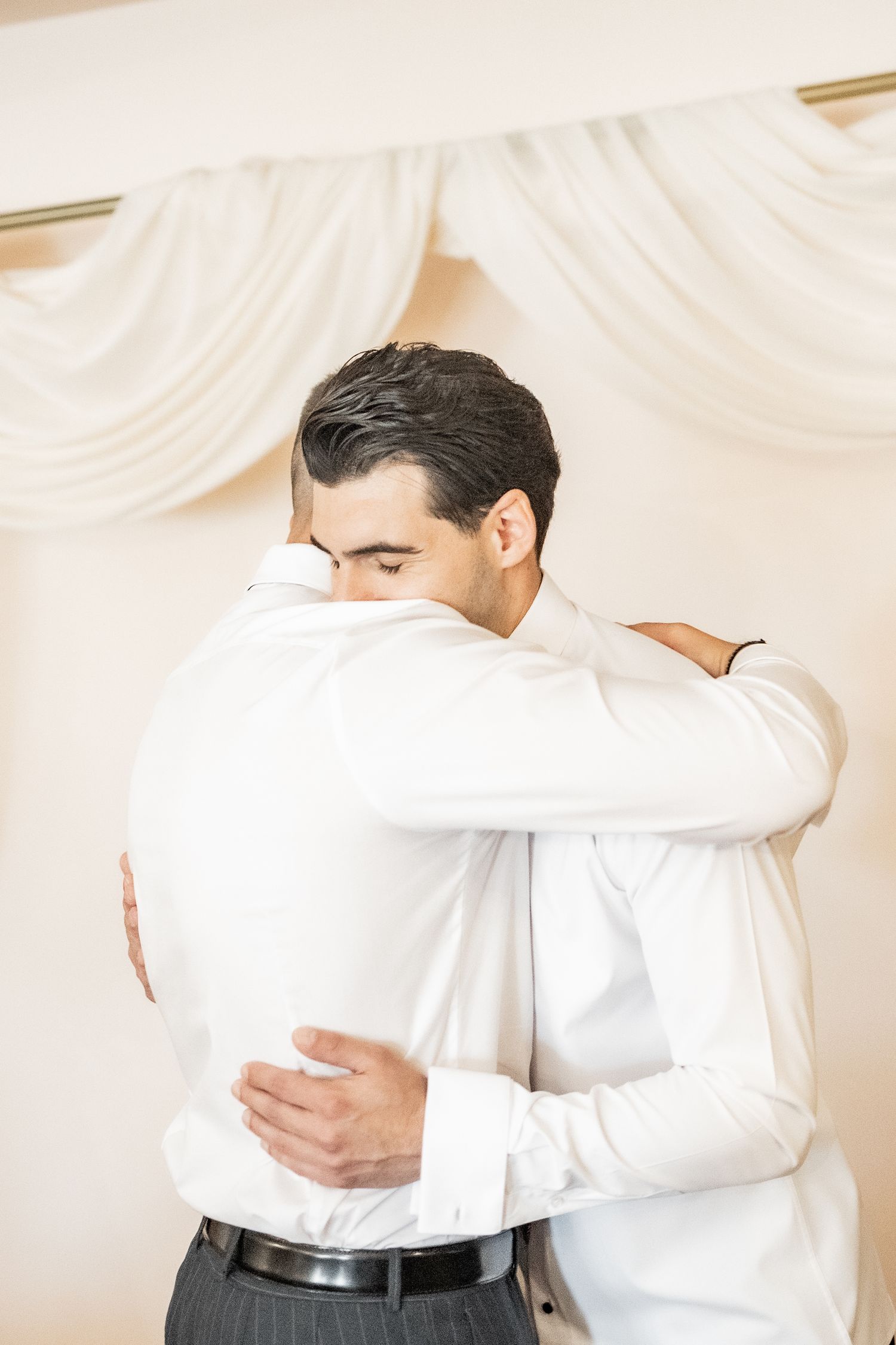 Two men in white shirts sharing a warm embrace at a formal event, standing near elegant draped curtains.