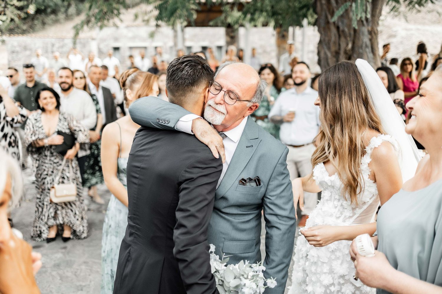 Wedding guests gather outdoors under trees for a celebration ceremony.