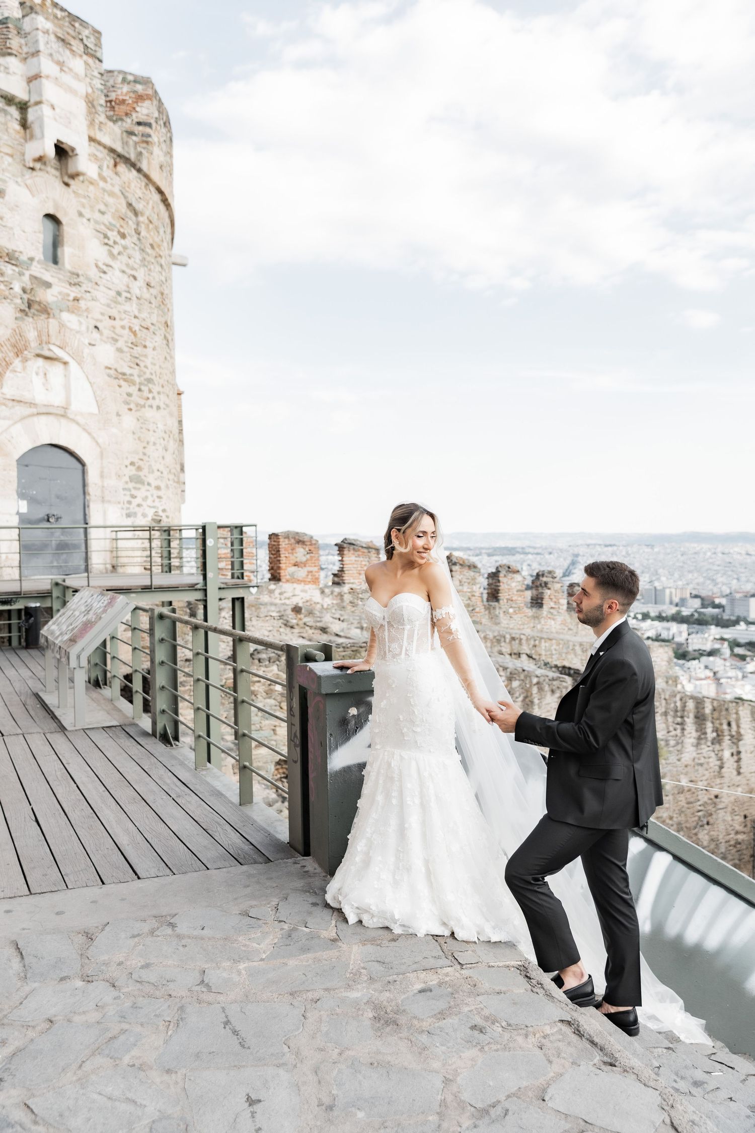 Bride and groom hold hands on a historic castle terrace with city views, bride in white gown, groom in black tuxedo.