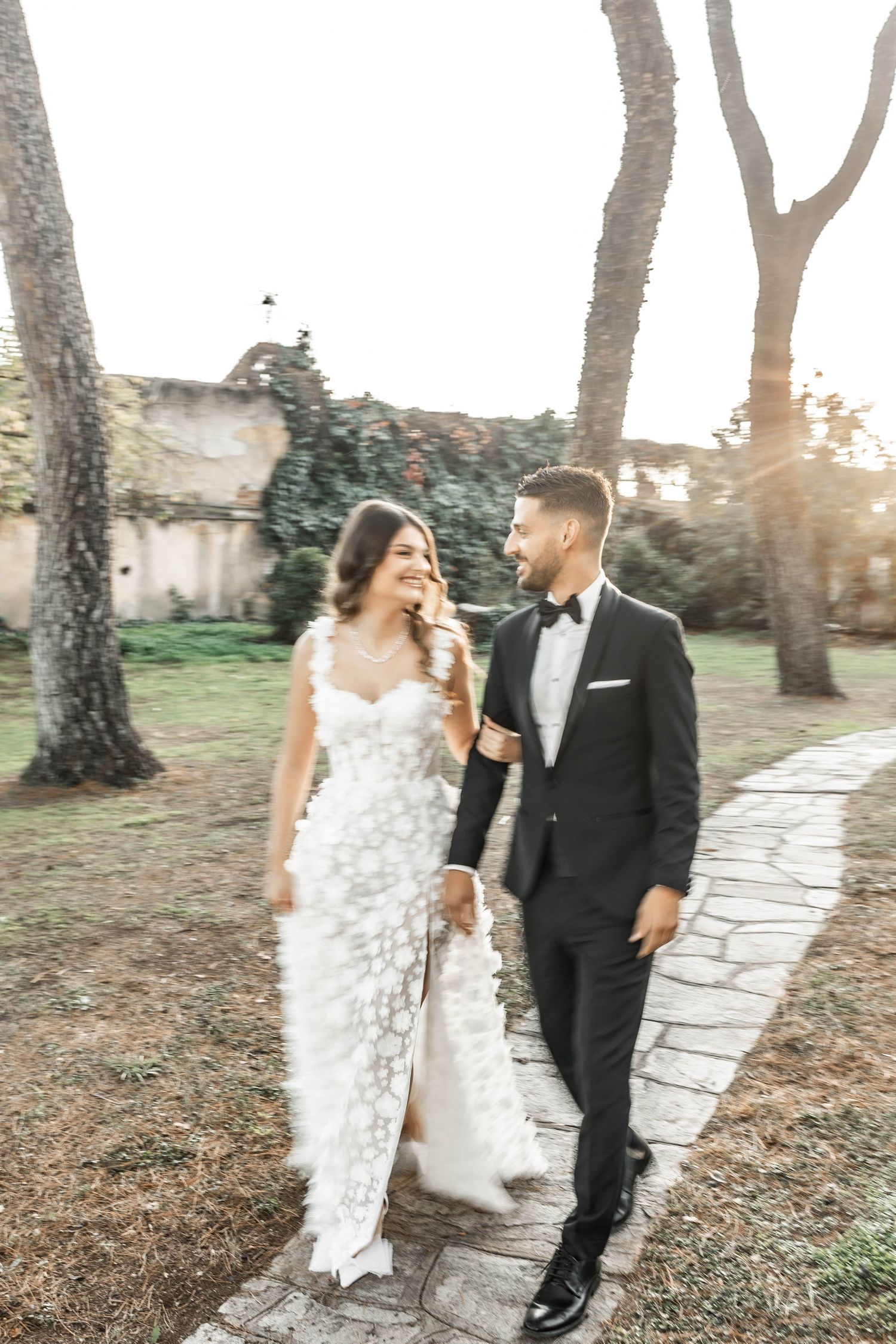 Couple walking along stone pathway surrounded by trees during golden hour.