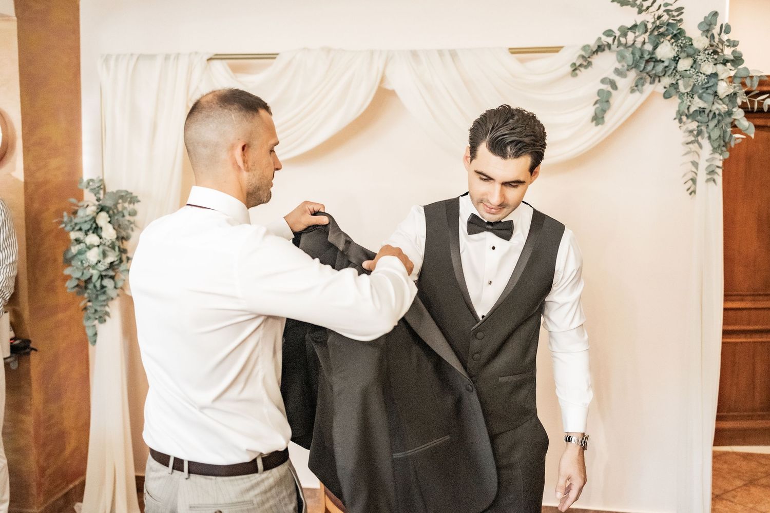Groomsman helping groom put on tuxedo jacket before wedding ceremony near floral arch backdrop.