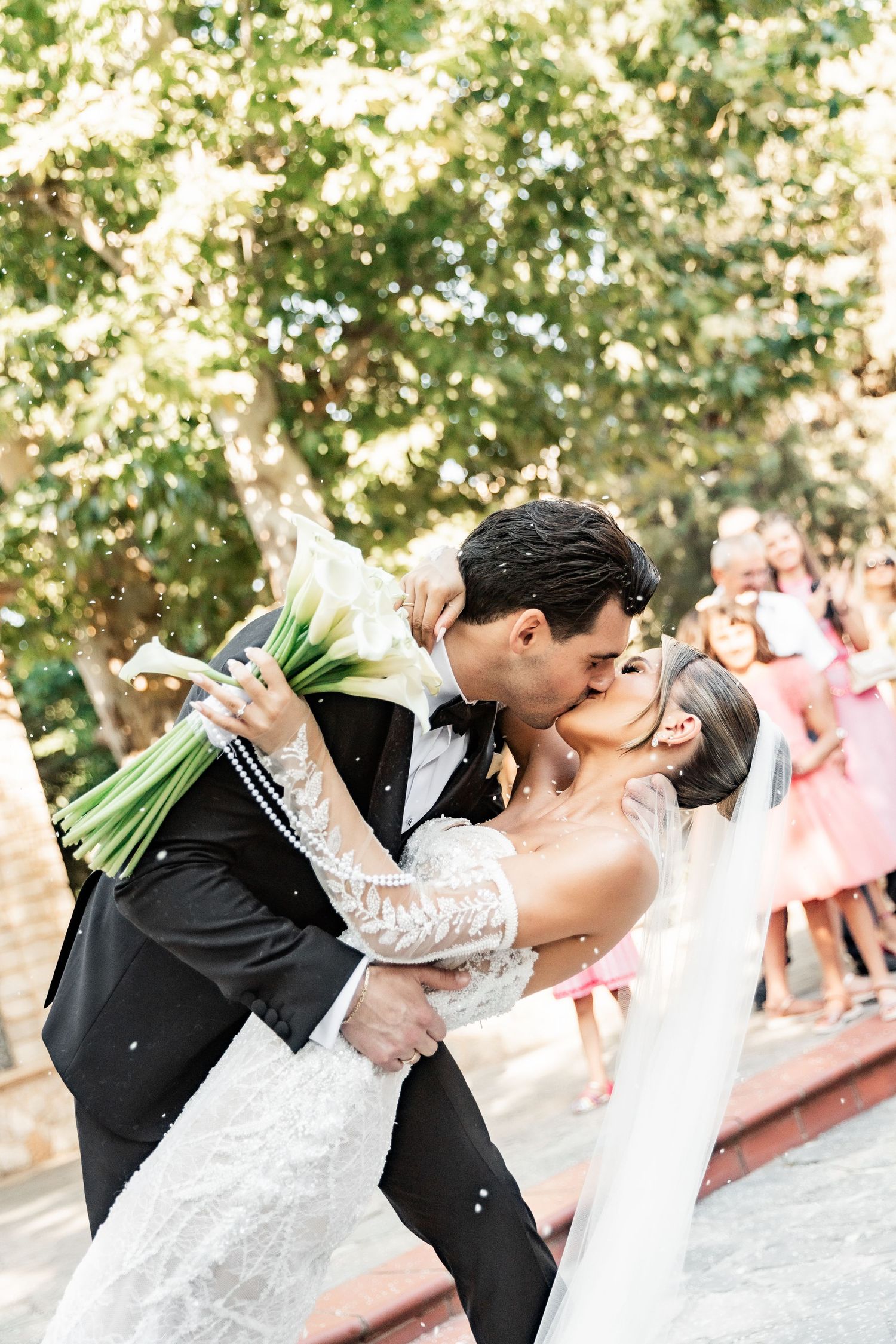 Bride and groom share a romantic dip kiss outdoors, with guests celebrating in the background at their wedding.