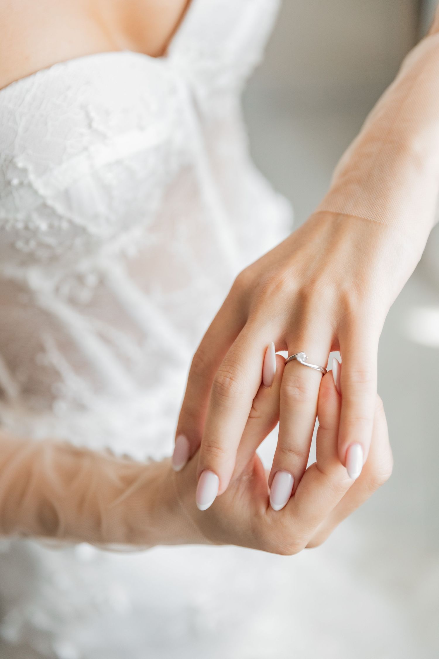 Bride in white lace dress displaying elegant diamond engagement ring on her finger with pink manicured nails.