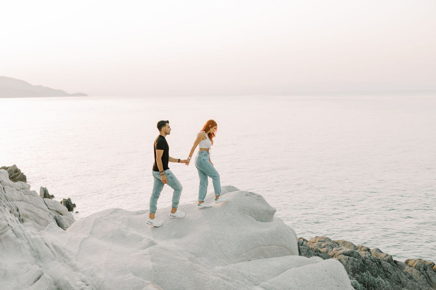 Two people explore a scenic rocky coastal area at sunset as waves crash against the shore.