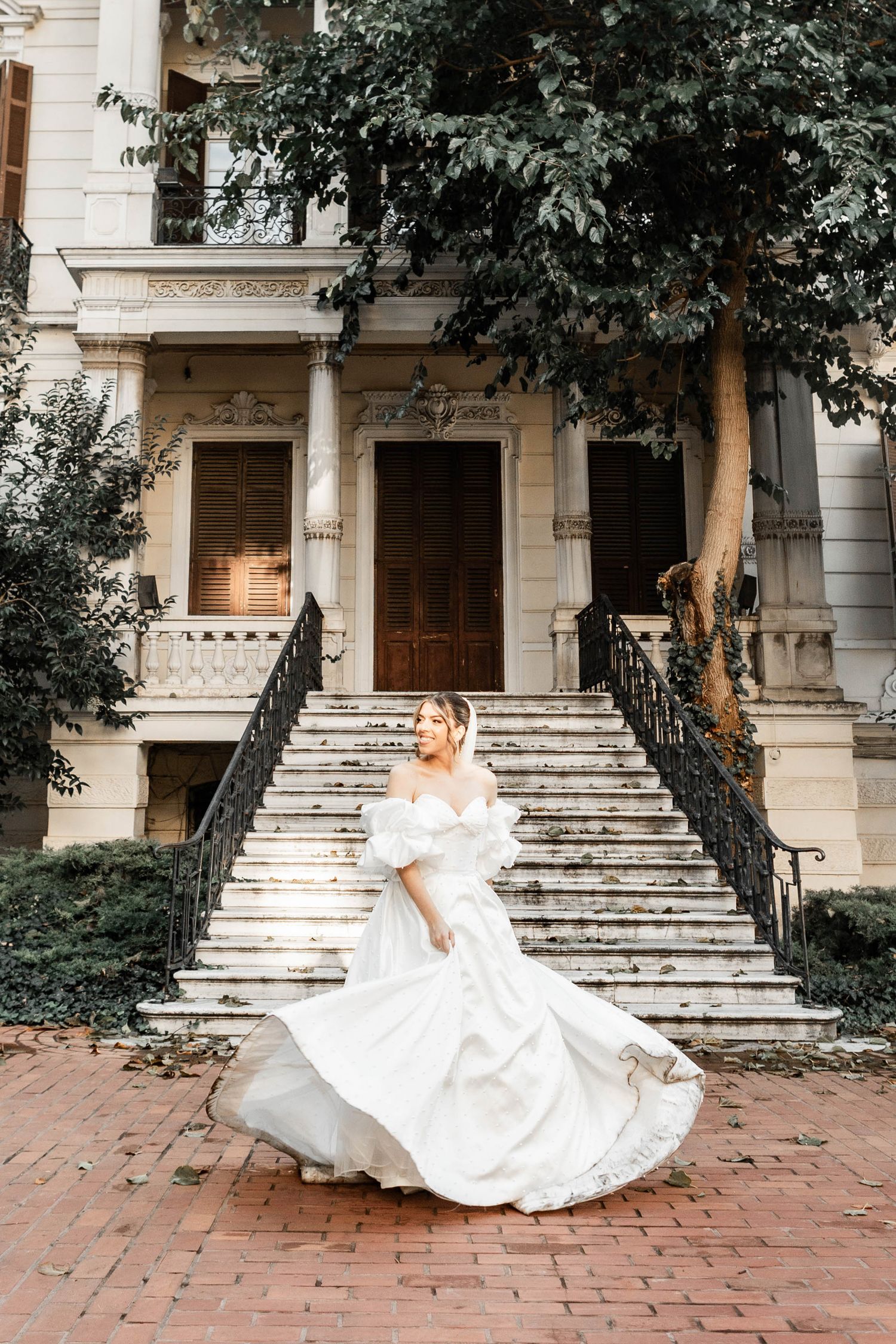 Bride in flowing white gown twirls on brick steps of a classic mansion with lush greenery.