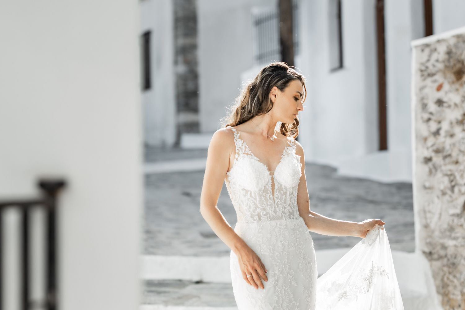 Elegant bride in fitted lace wedding dress poses against white Mediterranean building.