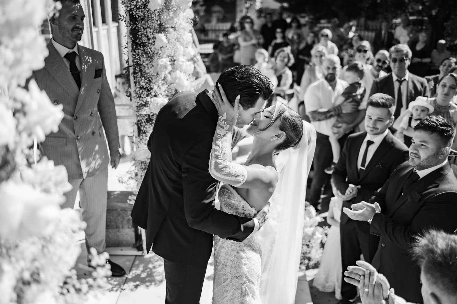 Black and white wedding photo of bride and groom sharing first kiss at outdoor ceremony with guests applauding.