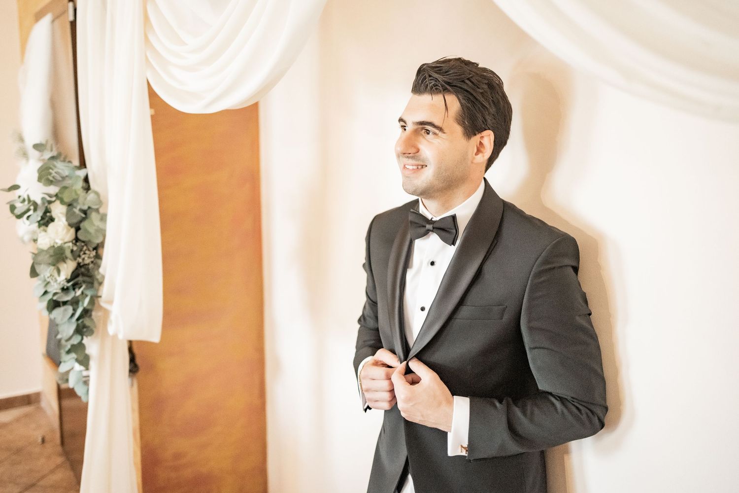 Groom in black tuxedo with bow tie buttoning jacket, smiling in elegant white draped wedding venue.