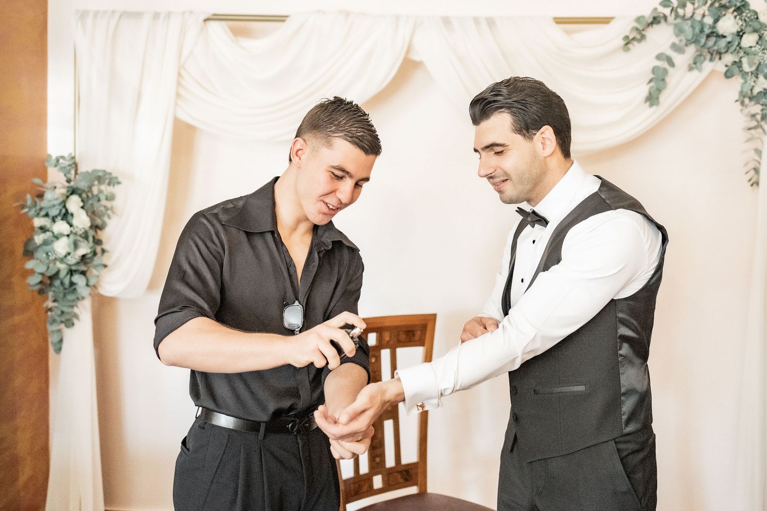 Two well-dressed men in formal attire examining a watch at a wedding venue with white drapes.