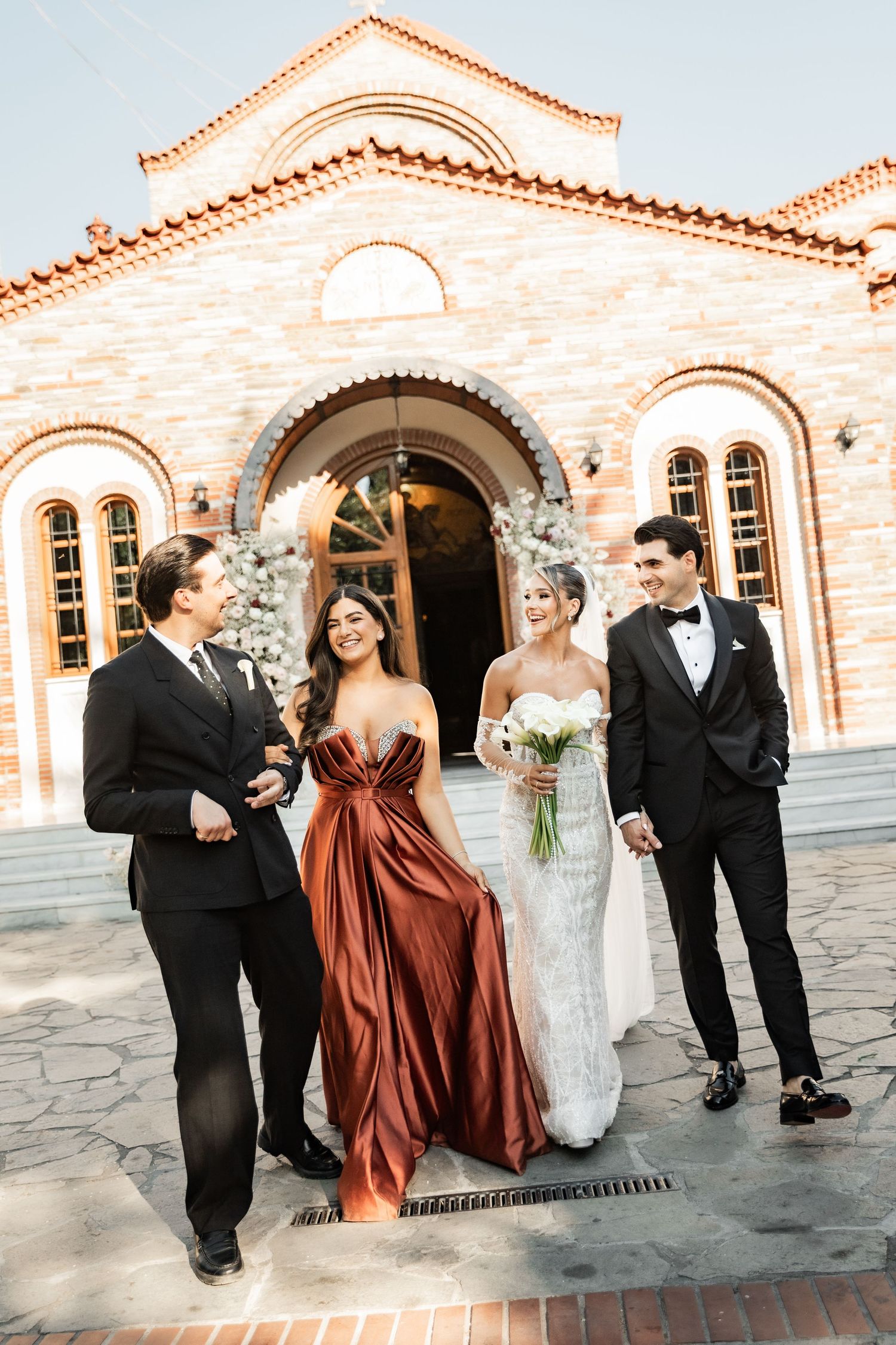 Wedding party of four posing outside a stone church, bride in white gown holding bouquet, bridesmaid in rust dress, groomsmen in tuxedos.